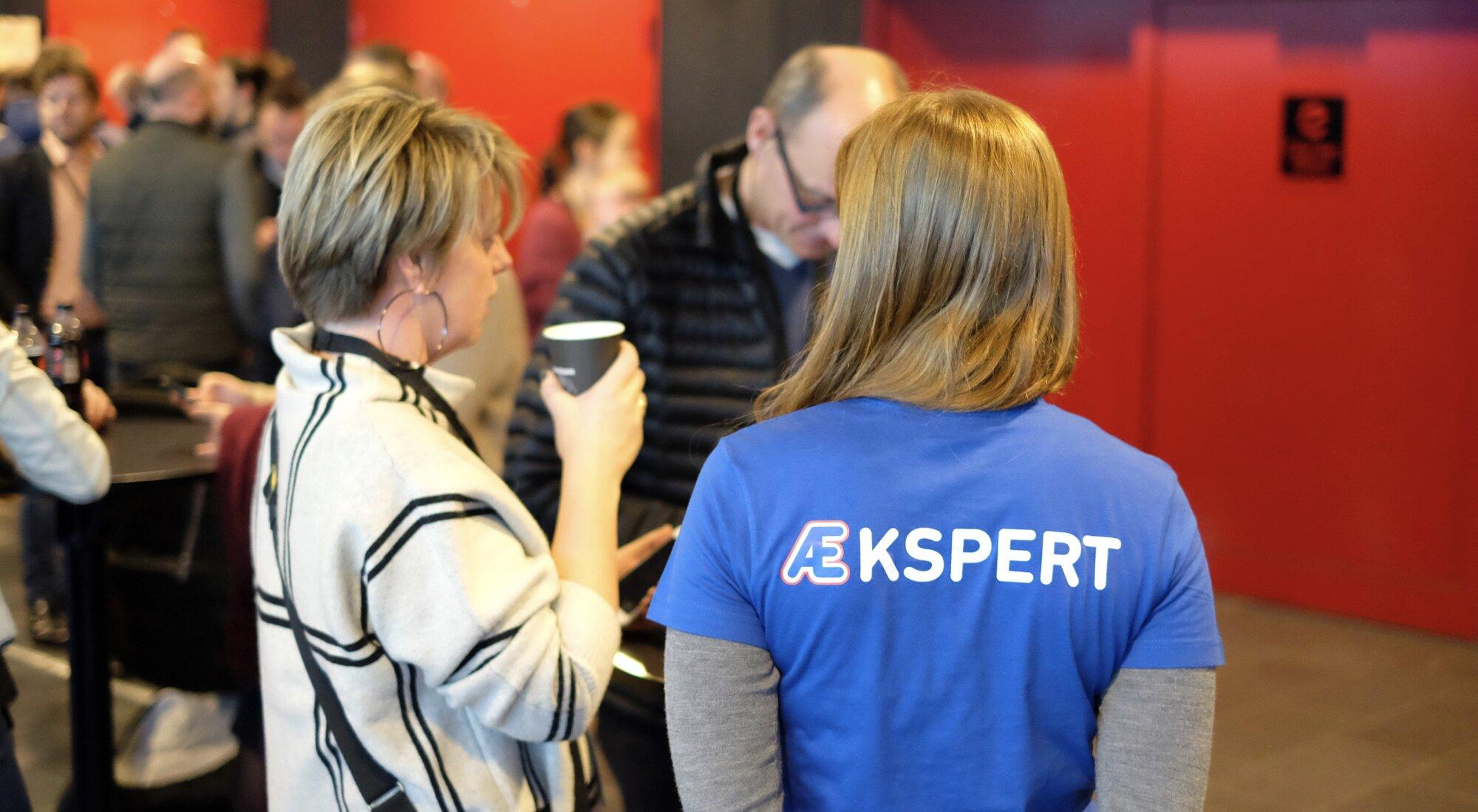 Grupper av mennesker i et rom. Fremst i bildet er en gruppe på tre, hvorav en står med ryggen til. Hun har på seg en blå t-skjorte med teksten "ÆKSPERT" påskrevet. Æ-en er uthevet i Rema1000s brandfarger.
