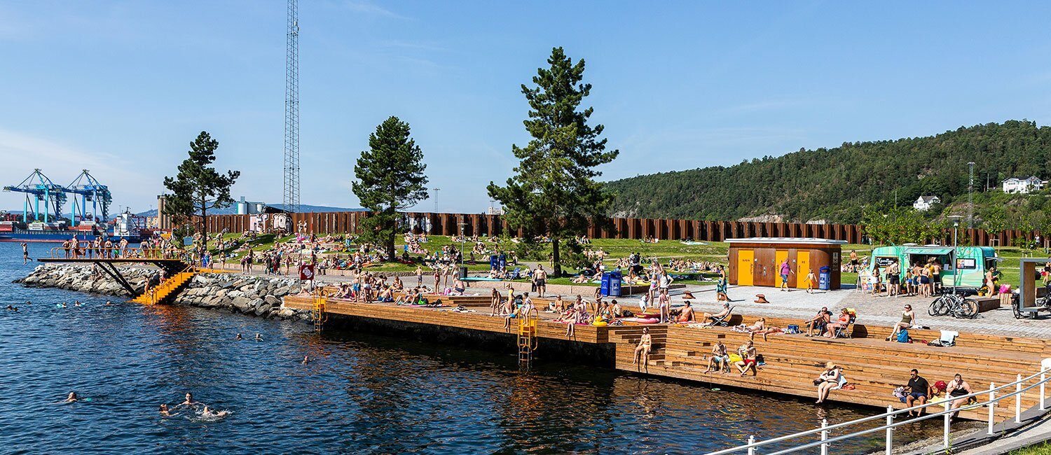 Bildet viser en badeplass med lang badebrygge på en sommerdag. Det er mange besøkende.