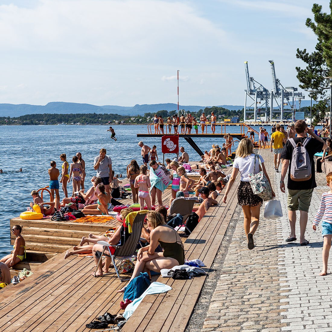 Bildet viser en badebrygge på en sommerdag. Det er mange folk, og litt bak i bildet mange som står på et stupebrett og venter på å få hoppe i vannet. I bakgrunnen ses Oslofjorden med et nes som stikker ut, og to store kraner i en vik.