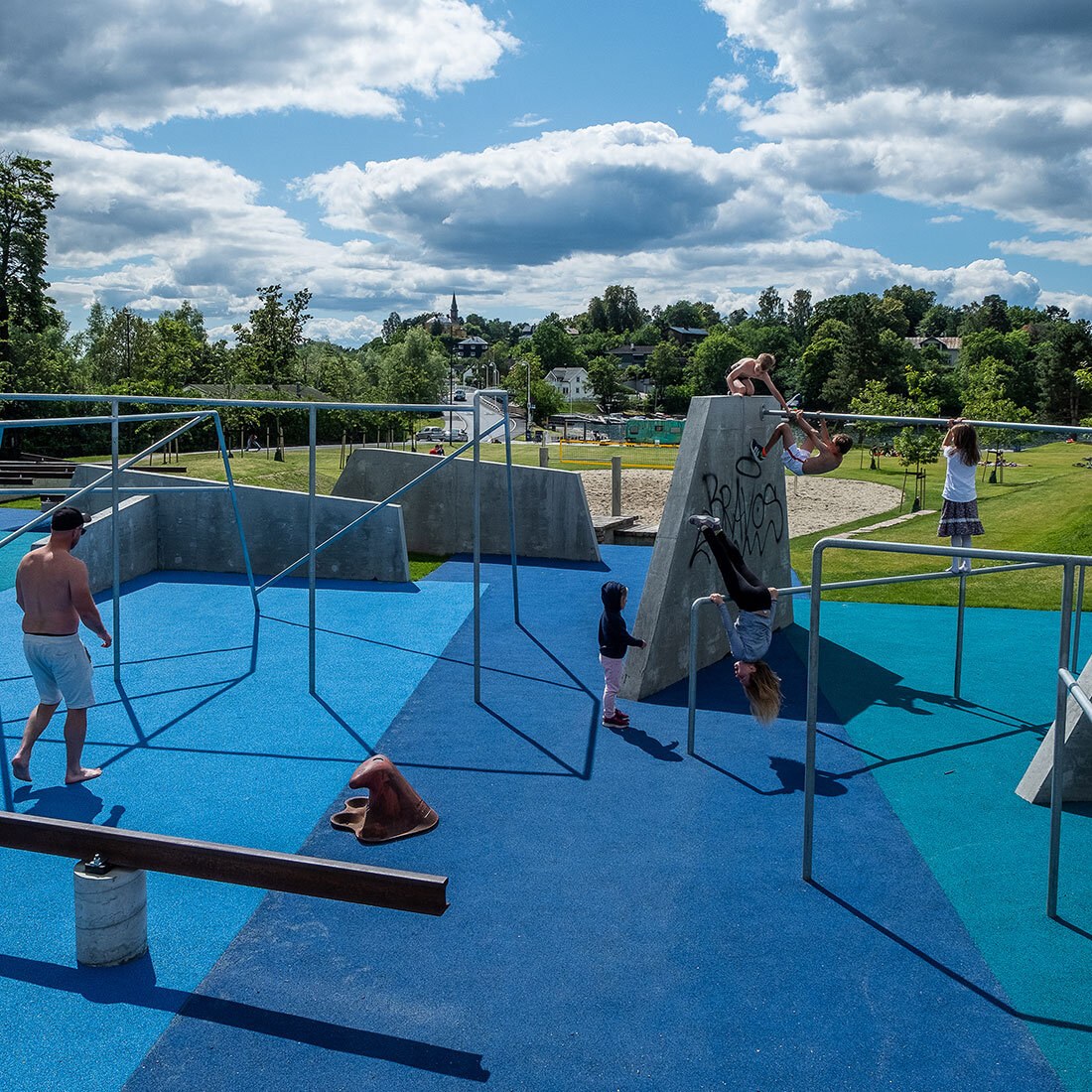 Et aktivitetsanlegg på en gummiert blått underlag. En håndfull store og små henger, klatrer og balanserer på stålrør og betongmoduler. I bakgrunnen ses mark og skog.
