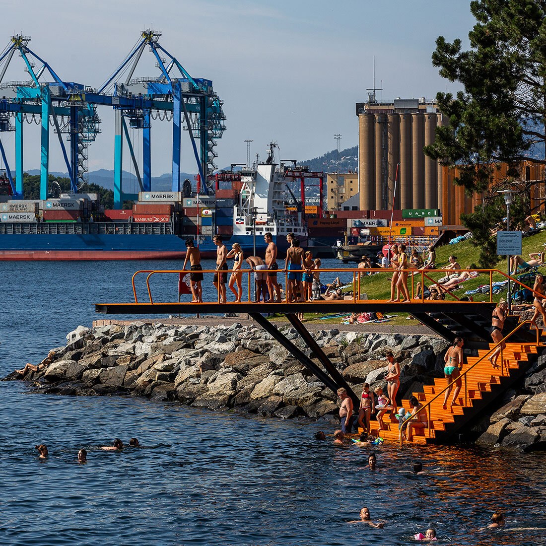 Bildet viser en kontrast mellom badende mennesker rundt et stort stupebrett, og industrielt landskap i bakgrunnen: store kraner, lasteskip, containere og et silobygg.
