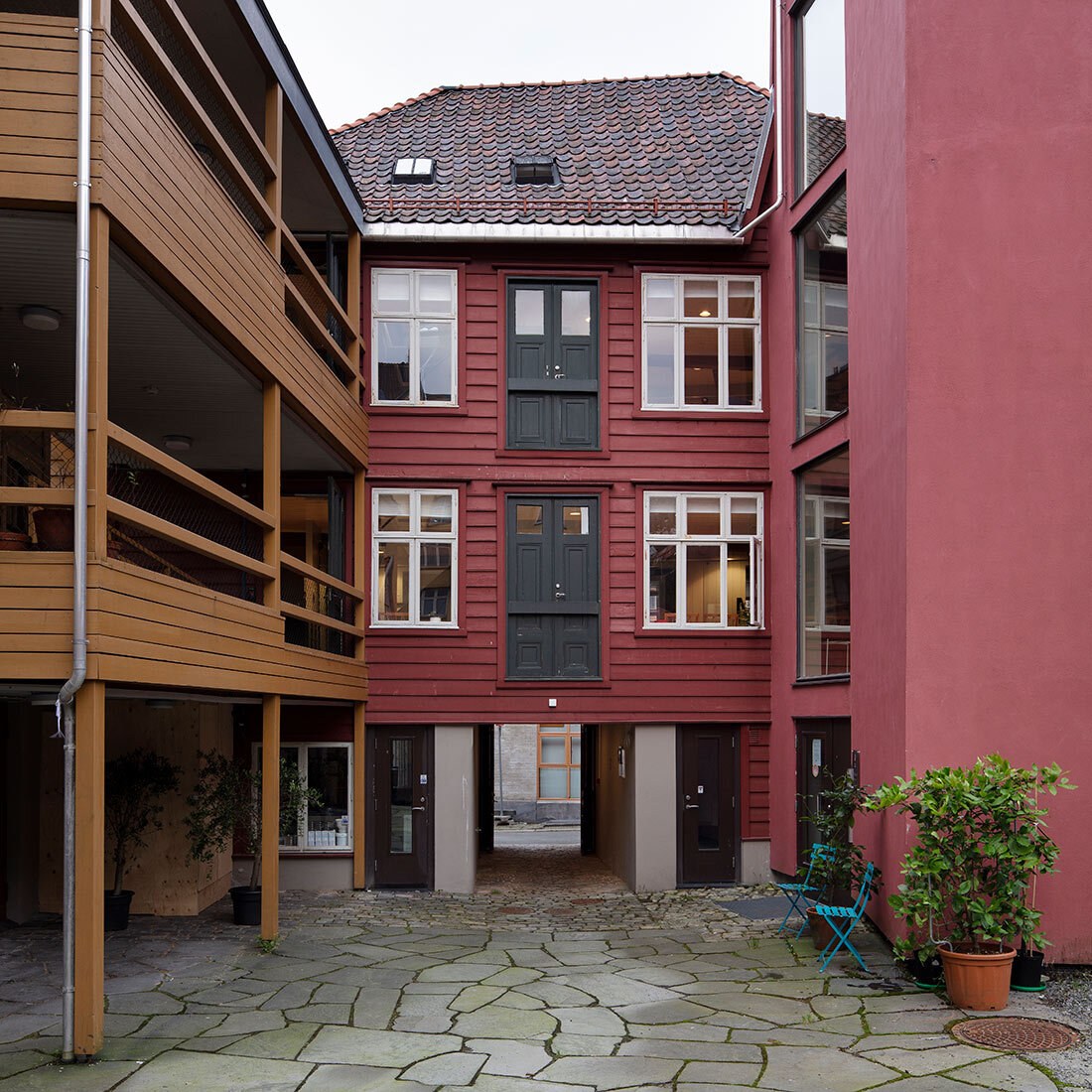 Et tredelt bygg sett fra en helledekt bakgård. sentralt i bildet er en panelkledd rødt hus over tre etasjer, i klassisk stil, med hvite vinduer med sprosser. Første etasje har et åpent inngangparti mot bakgården. Til venstre er et sennespgult bindingsverk som ser ut til å være av eldre dato, mens det til høyre i bildet er et moderne rødmalt påbygg i slett mur som føyer seg fint inn i resten av bebyggelsen.