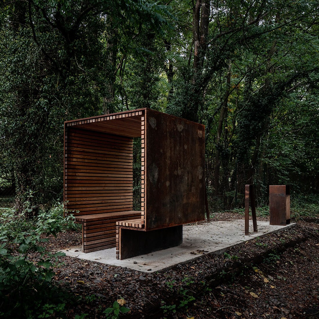 På en betongplatting i en skog ses et skulpturelt le-/sittemøbel med ramme av cortenstål og innside kledd i liggende trebjelker.