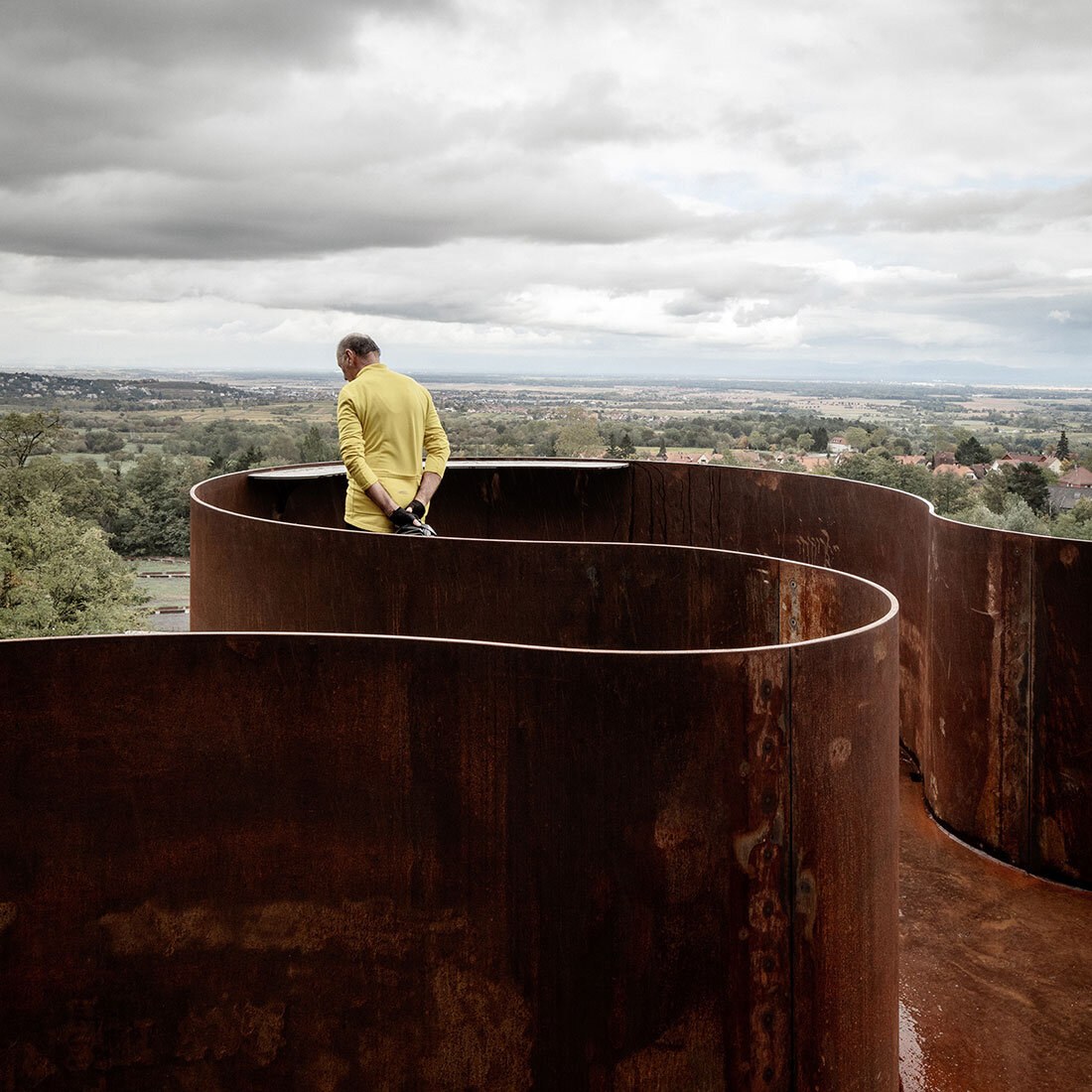 En mann i gul genser står på et utkikkspunkt utformet av buende cortenstål. Han har utsyn mot et landskap av spredt bebyggelse, åkrer og skog.