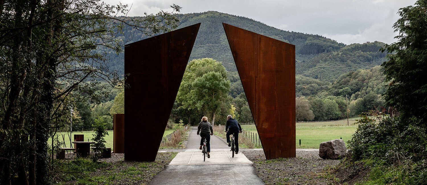To syklister ses bakfra mens de sykler på en landevei gjennom en slags portal ev to pilarer utført i cortenstål. Rommet mellom pilarene danner en trekant form mot himmelen. I bakgrunnen ses skogkledde åser.