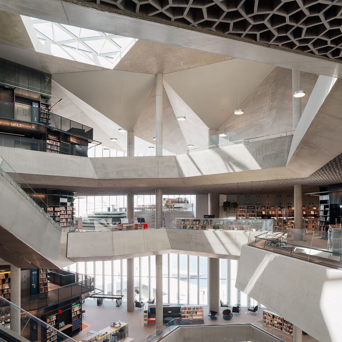 Moderne bibliotek med flere etasjer, åpne balkonger, betongkonstruksjoner og bokhyller, opplyst av dagslys fra et stort glasstak.