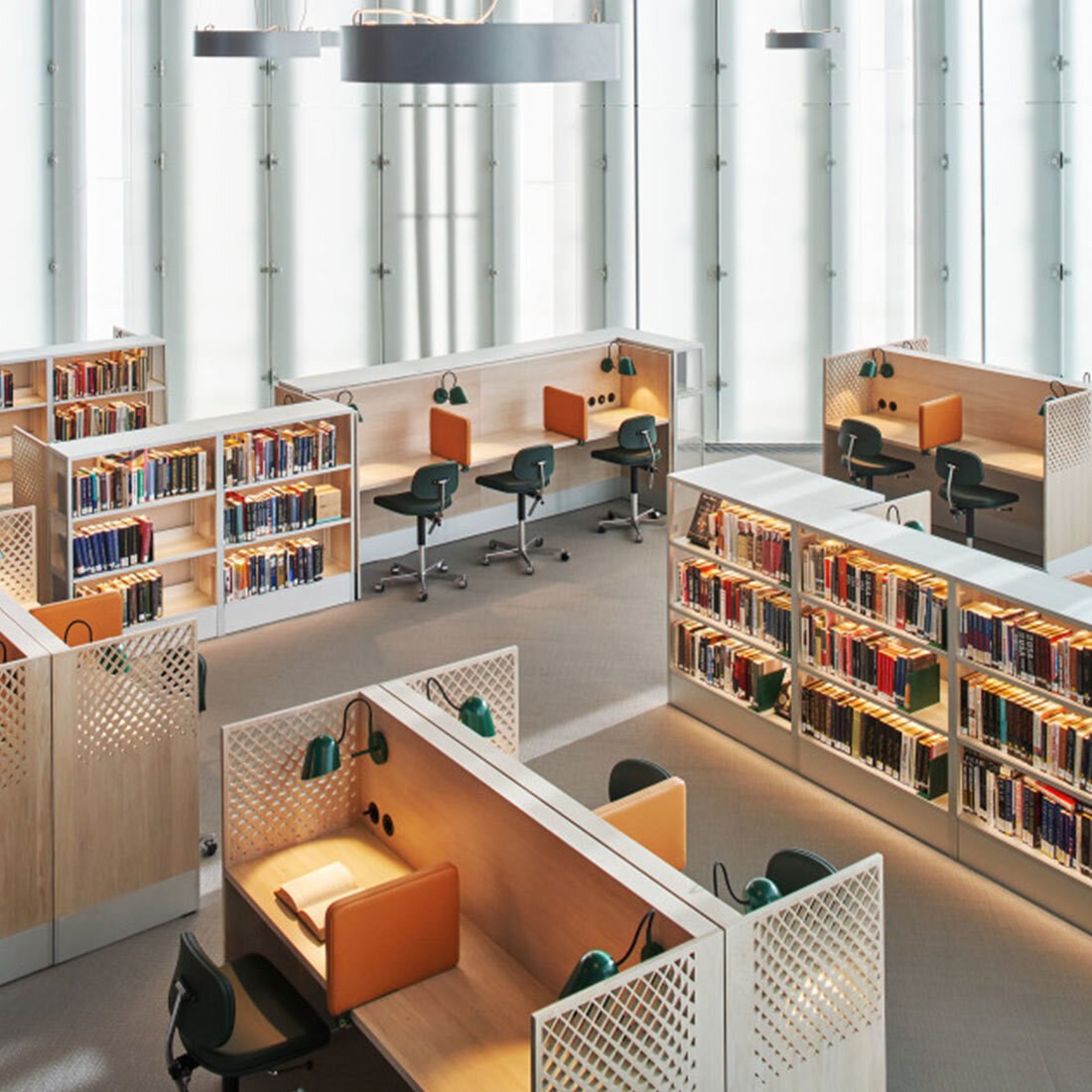 Leseplasser i et moderne bibliotek med avskjermede studiepulter, skrivebordslamper, kontorstoler og bokhyller fylt med bøker, i et lyst rom med store vinduer.
