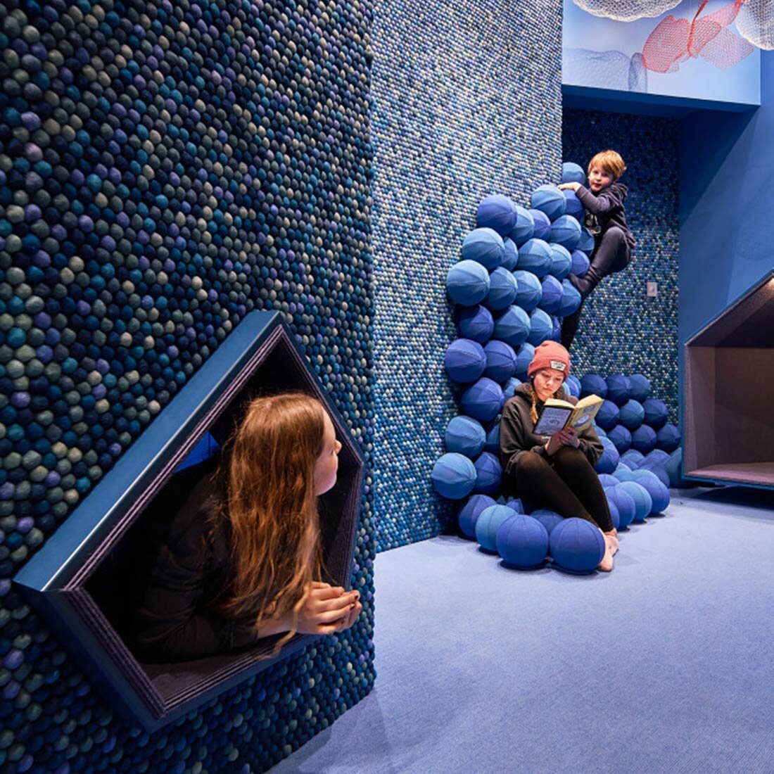 Et lekemiljø kledd i tekstil i ulike nyanser av blått. To barn leker og ett leser.