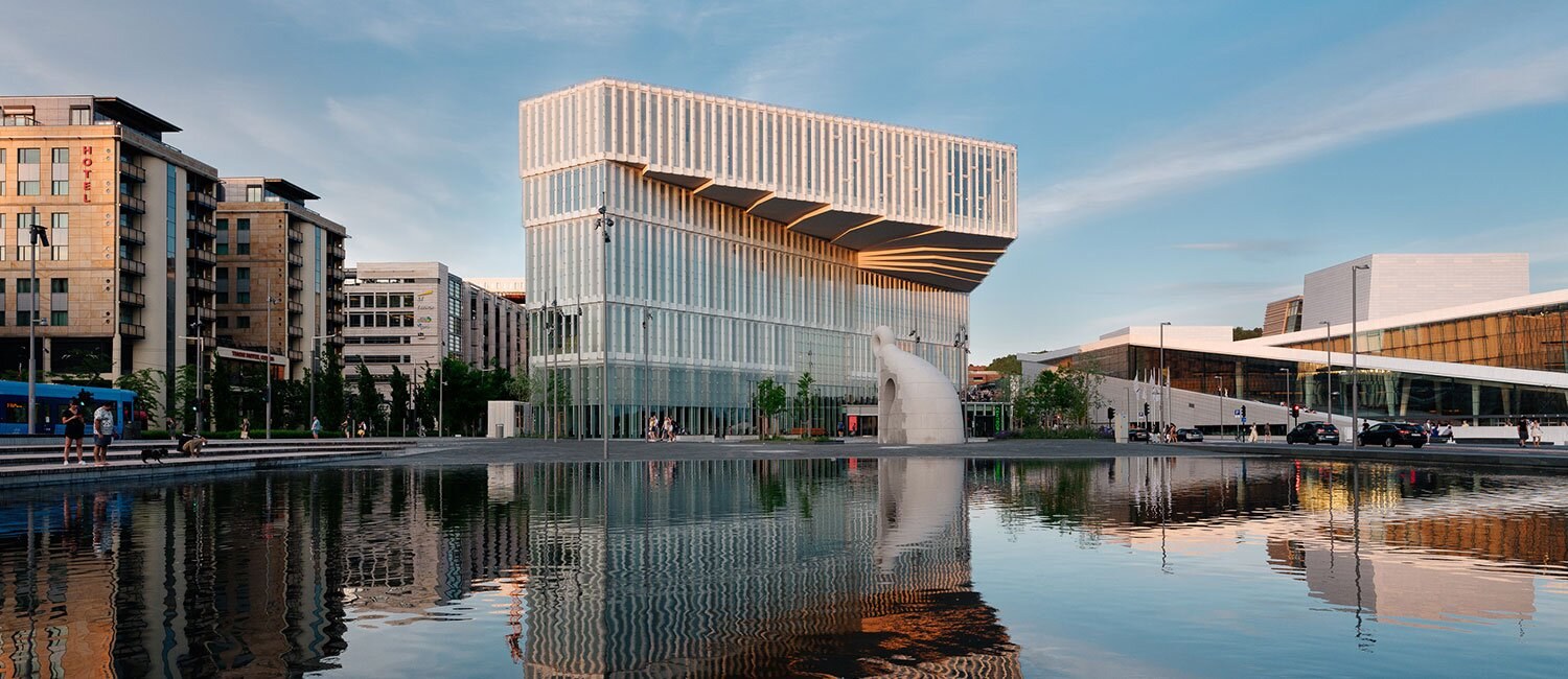 Fotografi av et særegent bygg i urban beliggenhet, men et vannspeil i forkant. Bygget er over ca fem etasjer, hvitt og vinduskledd. Det har et modernistisk uttrykk, og de to øverste etasjene er bygget utover byggets fotavtrykk på bakkeplan, i en slags organisk, lag-vis utforming. Bygget er ellers omgitt av mellomhøy kontorbebyggelse til venstre, og operaen i Oslo til høyre.