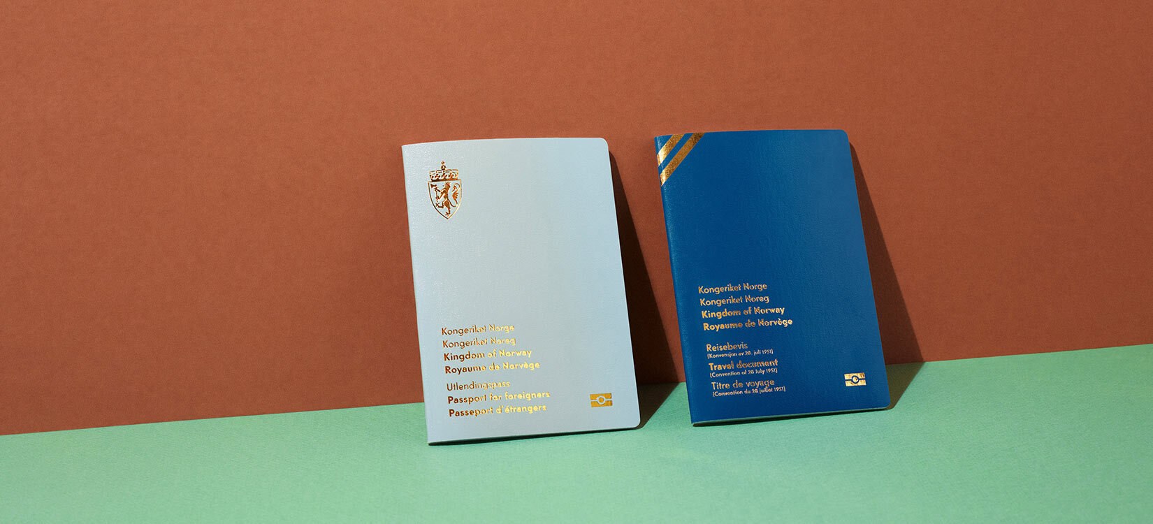 Et hvitt og et blått pass er stilt opp på et grønt bord med brun bakgrunn.