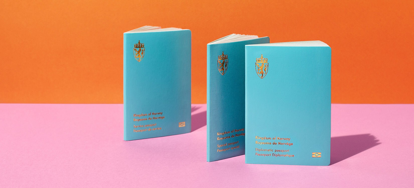 Tre pass med lyseblått omslag står oppstilt på et rosa bord med oransje bakgrunn.