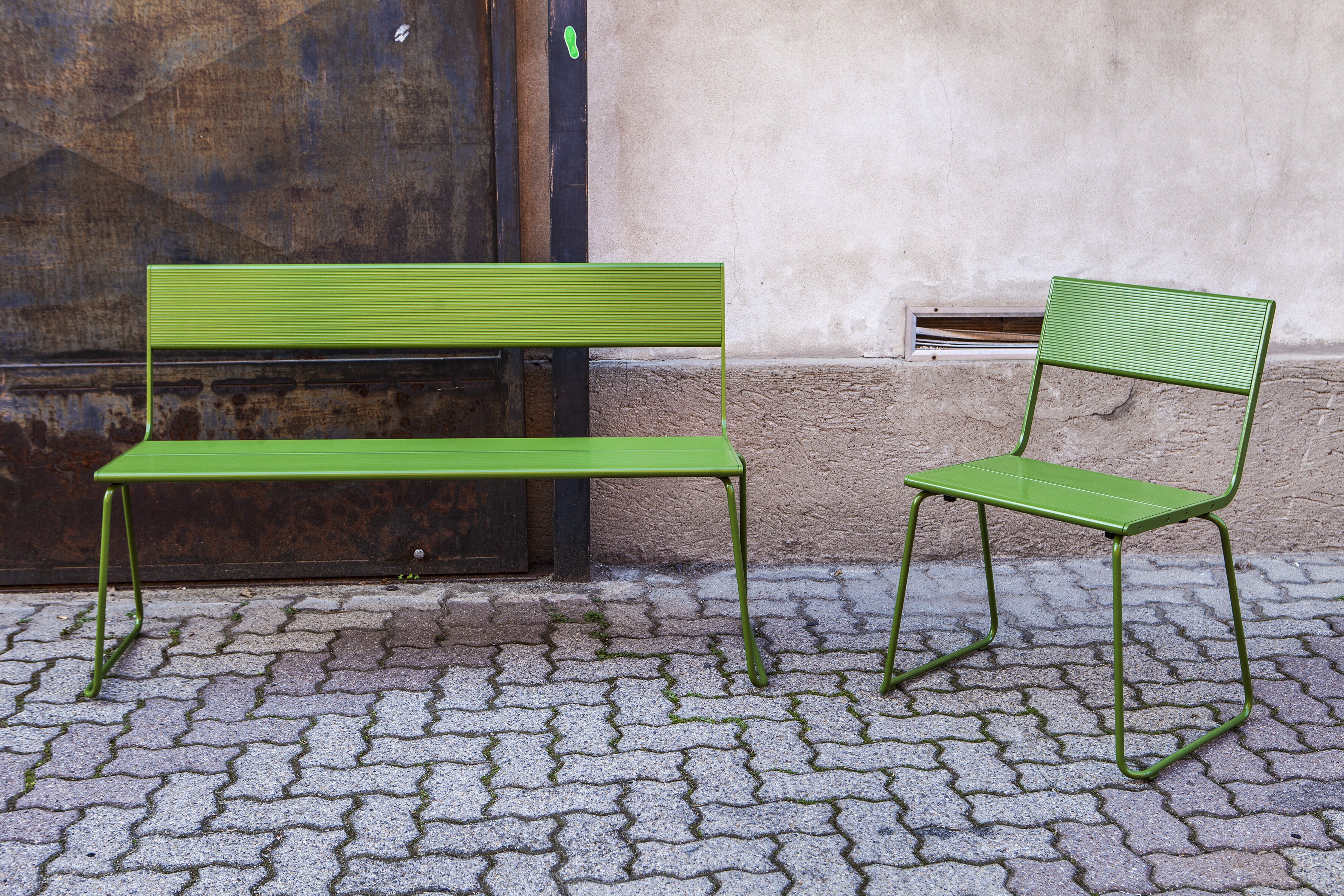 En benk og en stol plassert ute på mursteinskledd bakke. Møblene er utført i grønnlakkert metall.