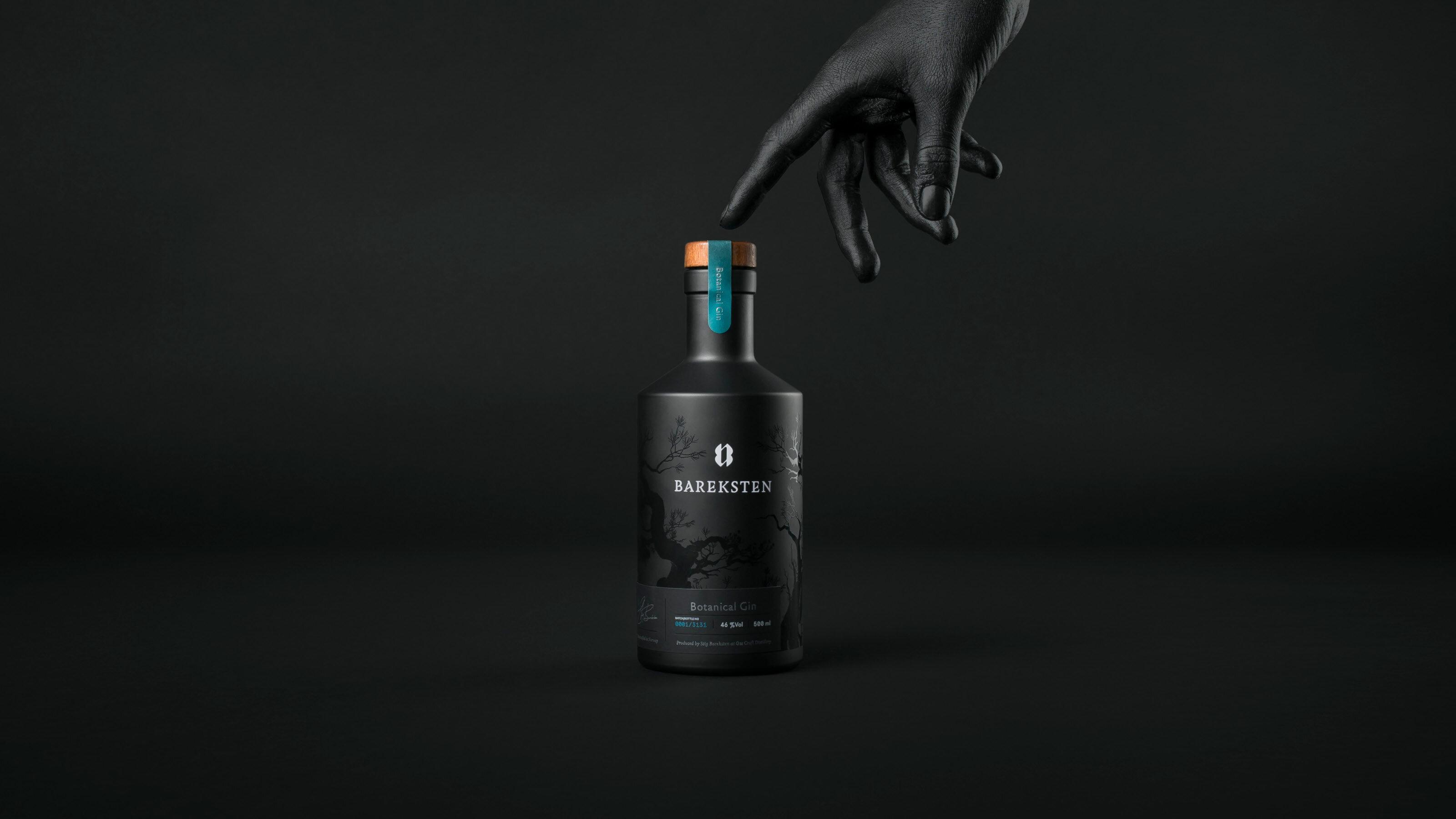 En svartmalt hånd peker på en svart brennevinsflaske på svart bakgrunn.