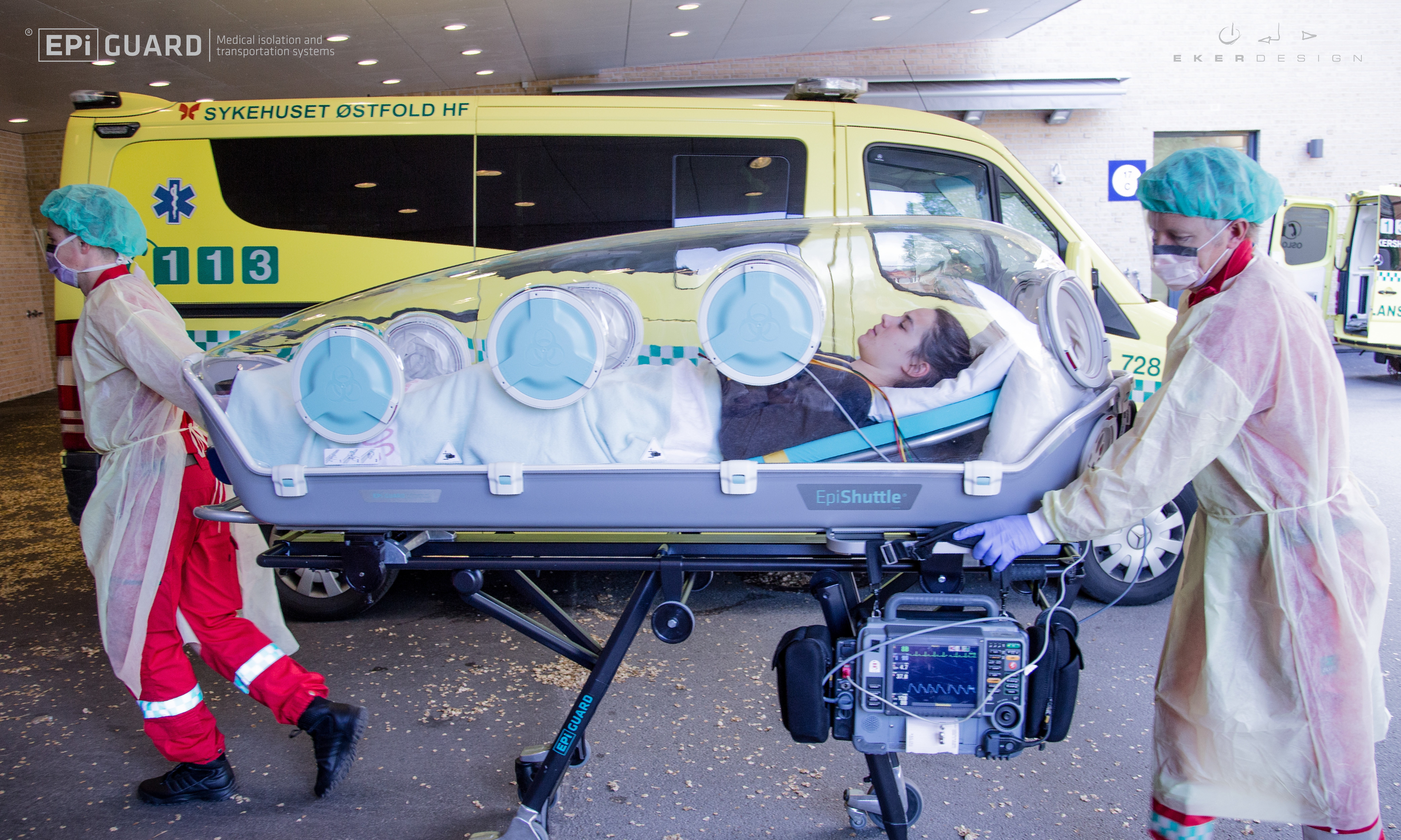 En pasient fraktes i en luftett båre som ligner en kuvøse. To ambulansearbeidere ruller båren ikledd smittevernutstyr.