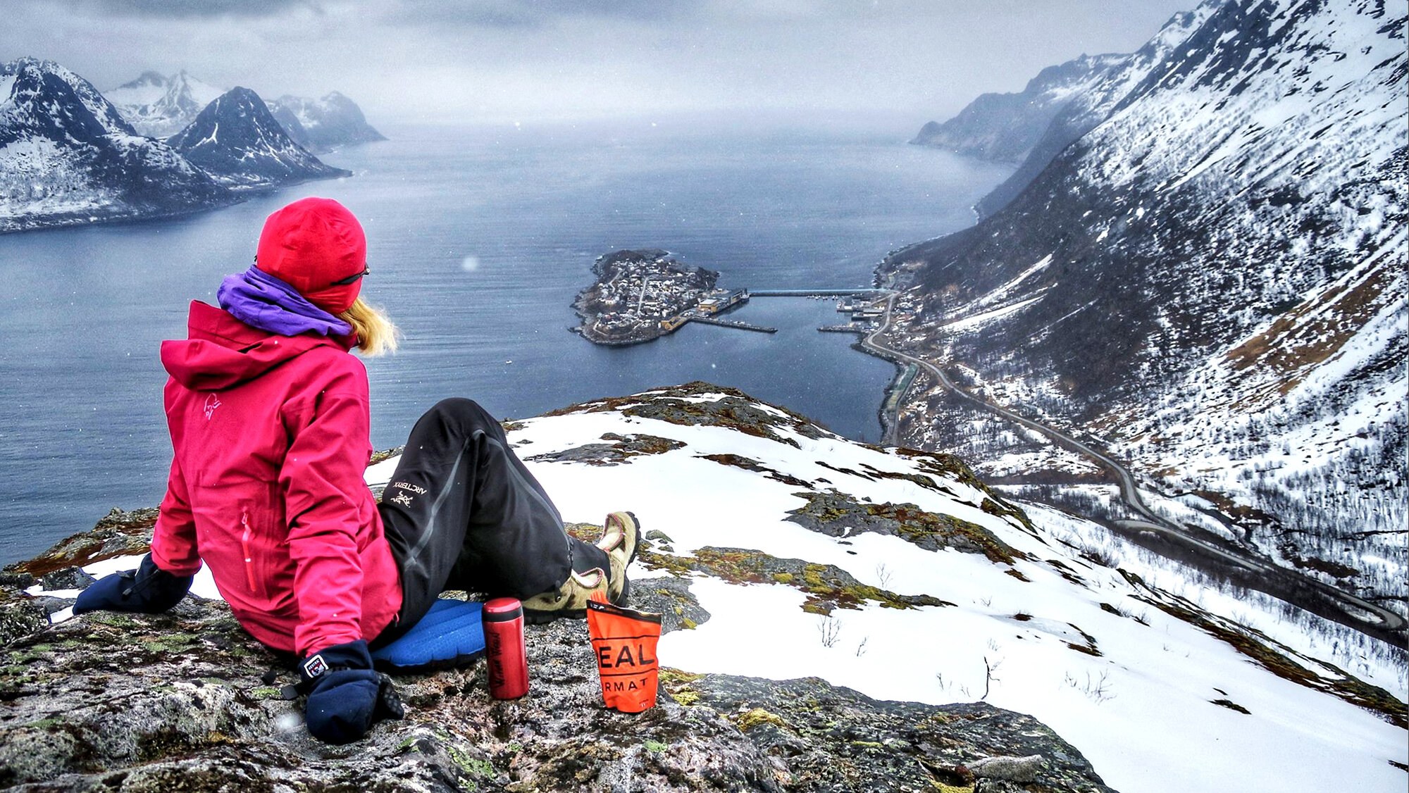 Bildet viser en kvinne sittende på en fjelltop som ser utover et fjordlandskap med hvitkledde fjelltopper. Ved siden av seg har hun en termos og en pose real turmat.
