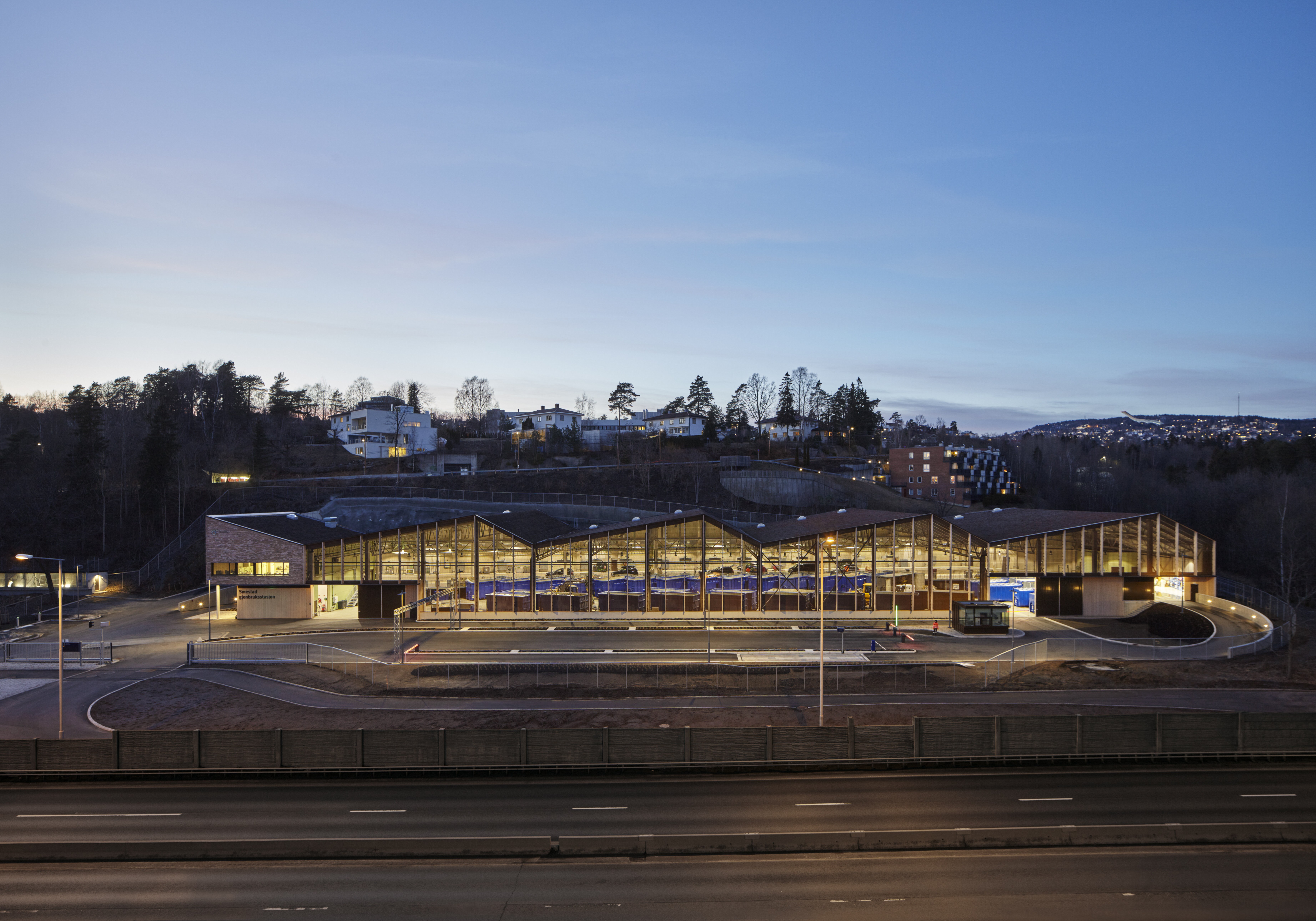 Oversiktsbilde over gjenbruksstasjonen. Den har en lav profil med karakteristisk vinklede takformer. Interiører lyser opp i skumringen.