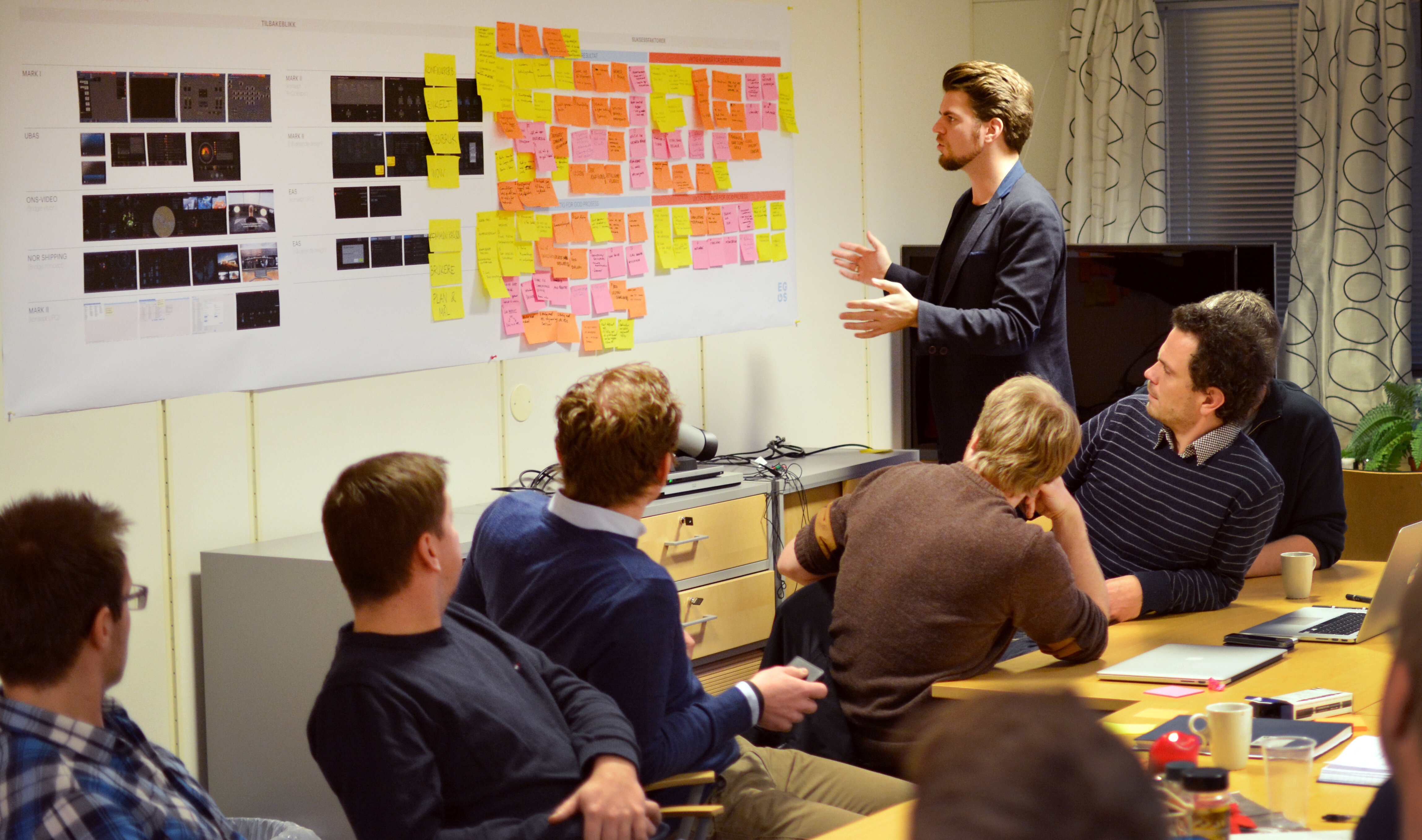 En forsamling i et konferanserom. En person står og gestikulerer mot et oppslag på veggen med post it-lapper, mens resten sitter, snudd mot ham.