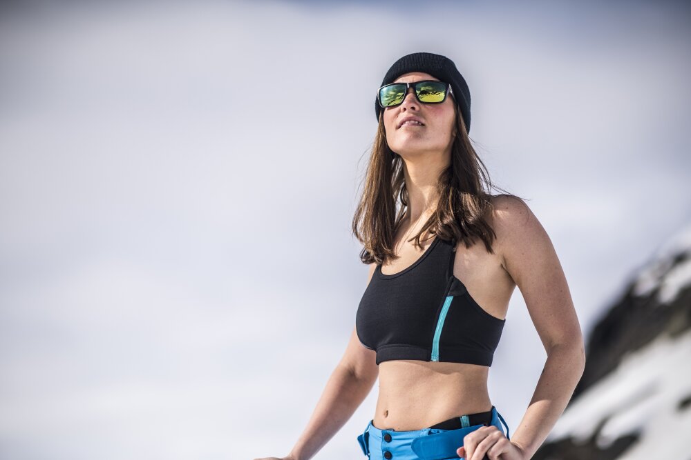 Halvnært bilde av en kvinne med solbriller og overkropp kun iført en trenings-bh med en glidelås i siden.