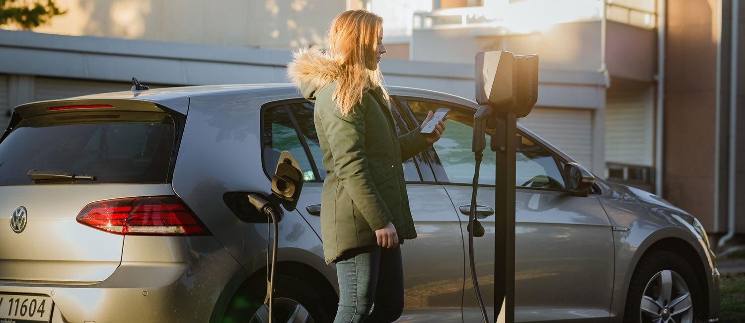 En kvinne står avslappet ved en ladeboks, mens en bil lader bak henne. Hun ser på telefonen.