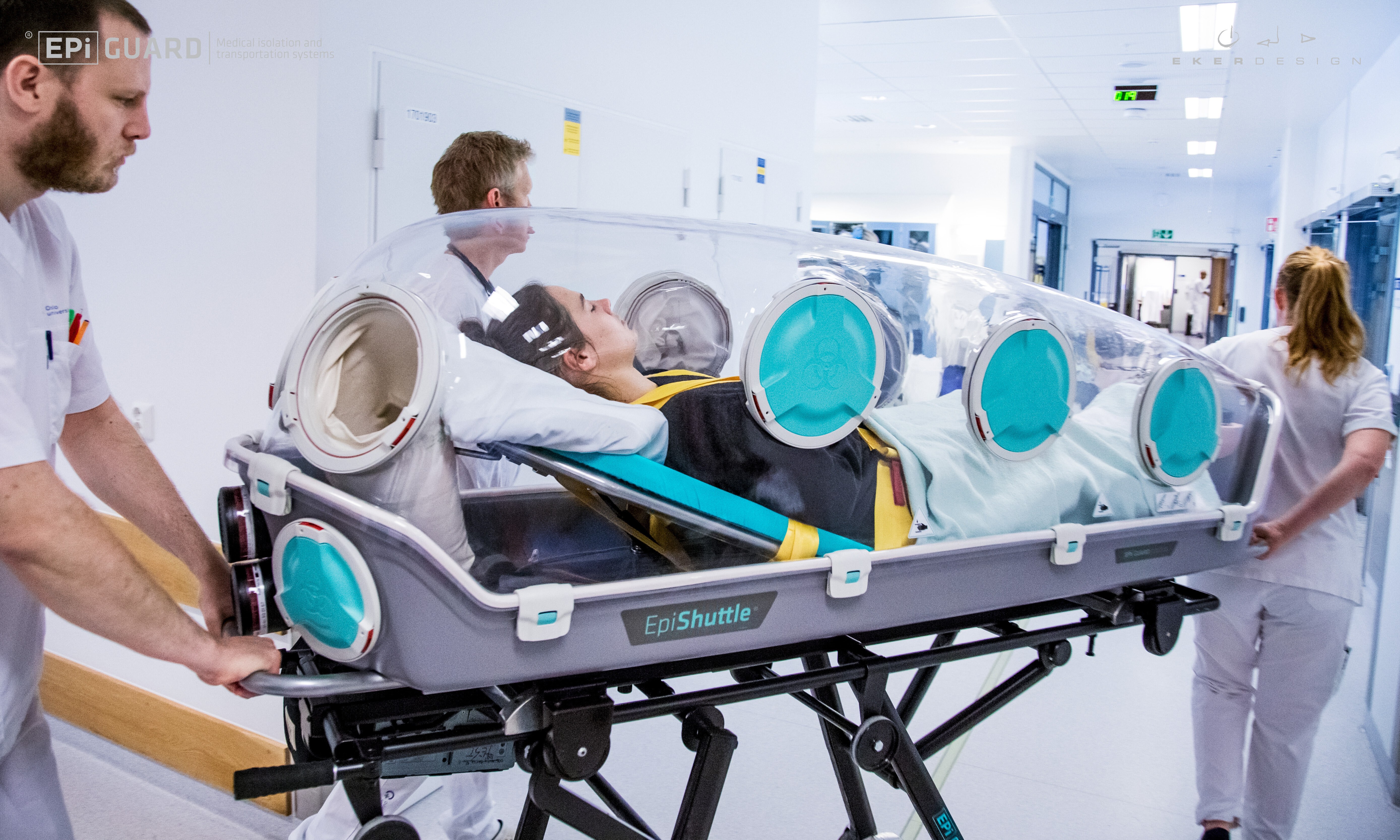 En båre med overbygging – som en kuvøse – trilles av tre hvitkledde sykepleiere gjennom en sykehuskorridor.