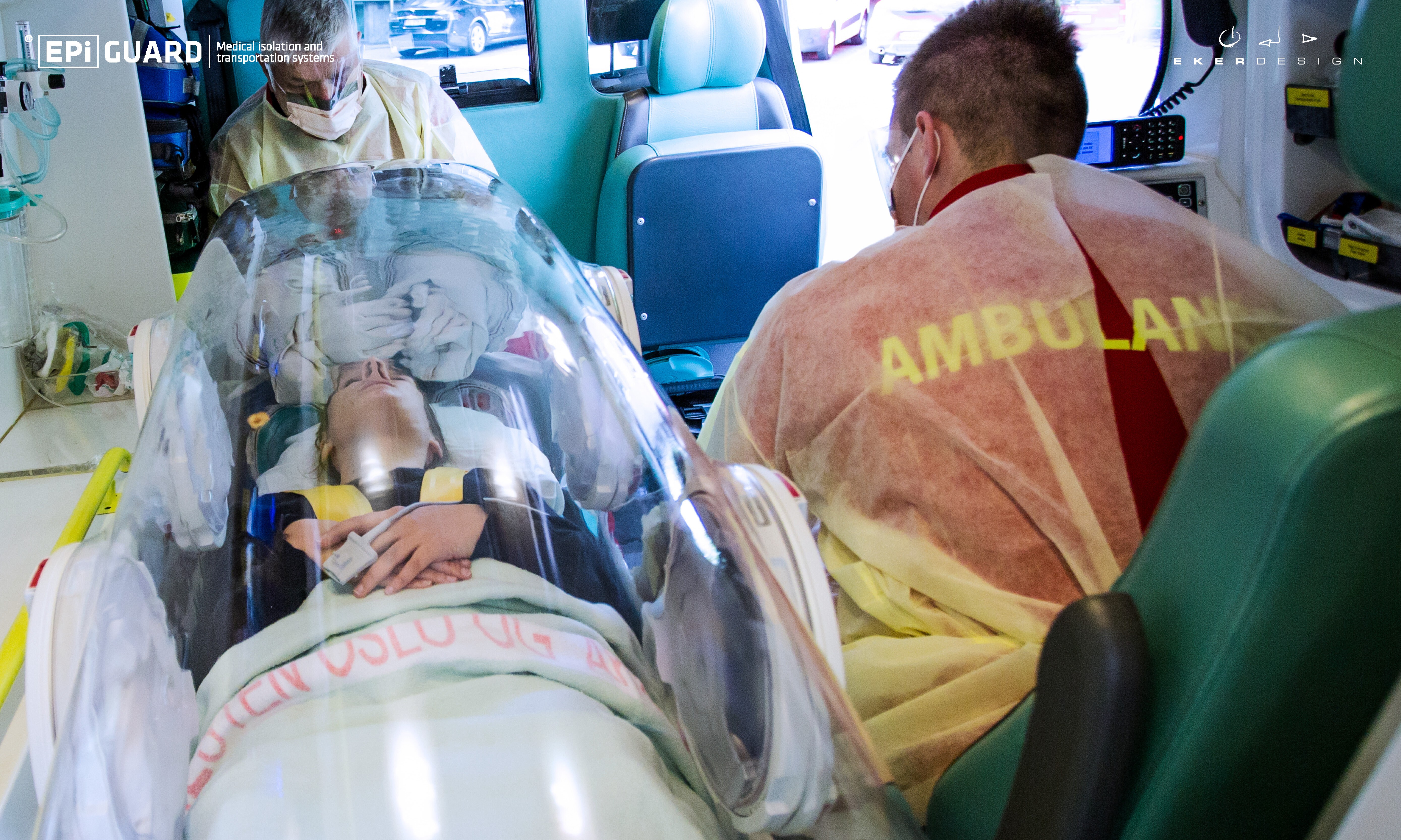 Foto fra innsiden av en ambulanse. En pasient er i en overbygget båre, mens to ambulansearbeidere i smittevernutstyr sitter på hvert sitt bilsete i hver ende av pasienten.