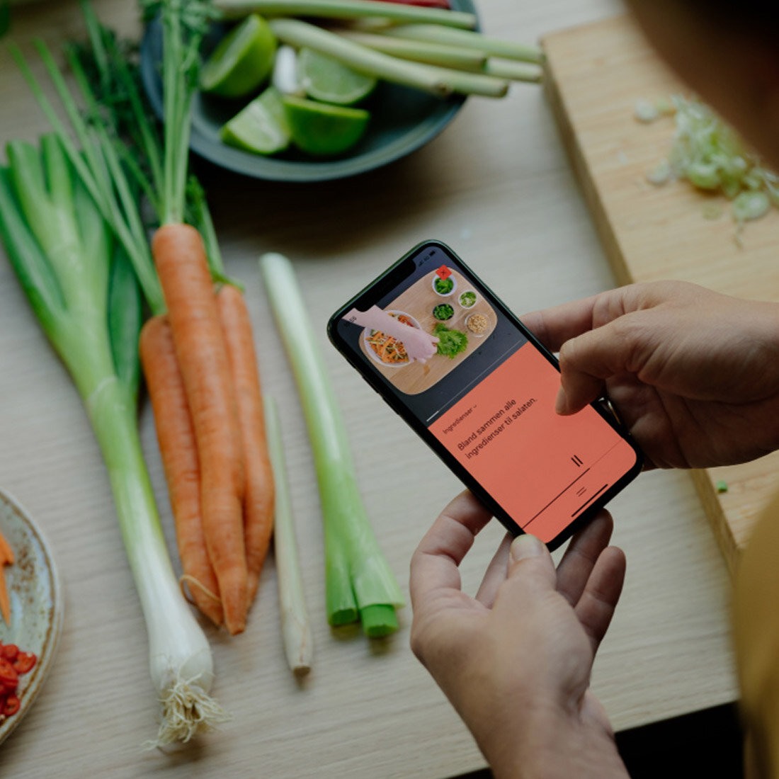 Fotografi av hender som holder en smarttelefon. På skjermen ses instrukser for matlaging. Telefonen holdes over en kjøkkenbenk dekt av grønnsaker og et skjærebrett.