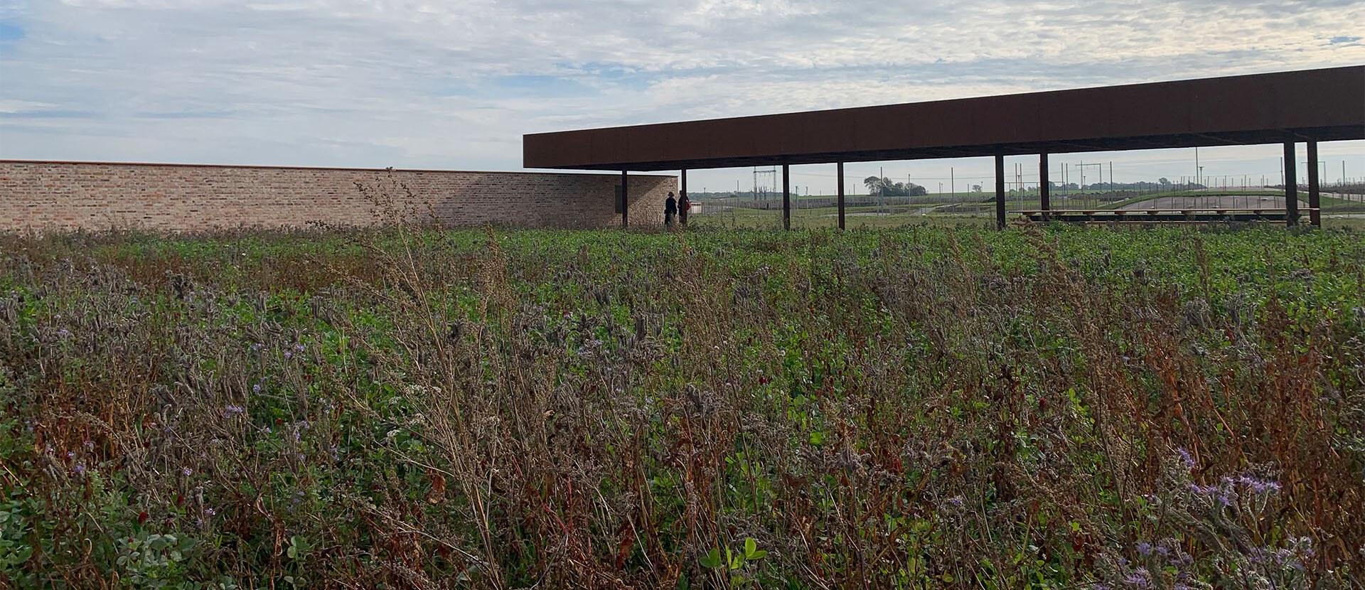 Fra et frodig jorde ses et monument bestående av to konstruksjoner: en teglsteinsvegg til venstre som møter et overbygg i mørkt metall.