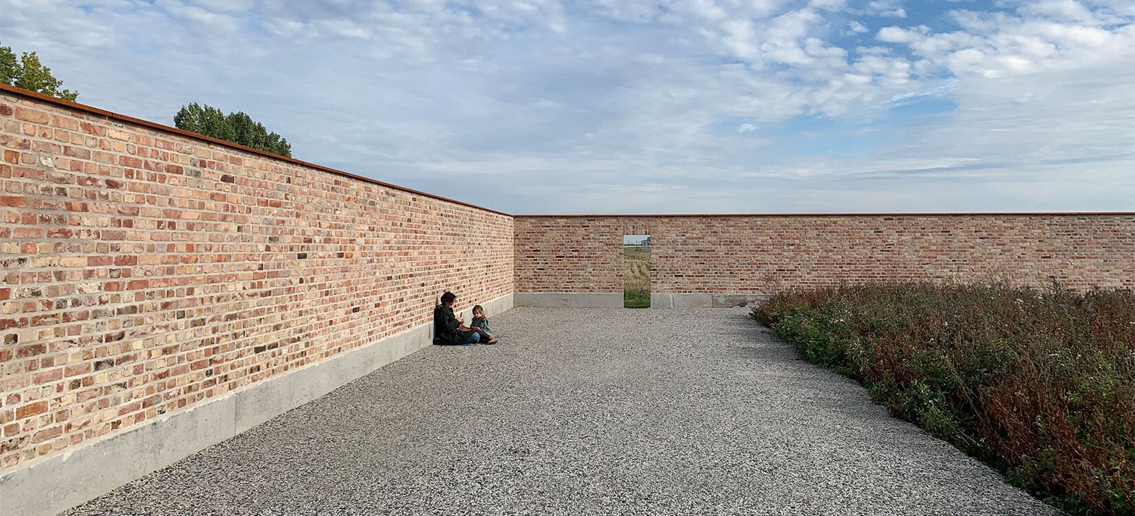En voksen og et barn sitter på grusen med ryggen mot en teglsteinsmur som omringer et frodig jorde. I ene enden av muren er åpning.