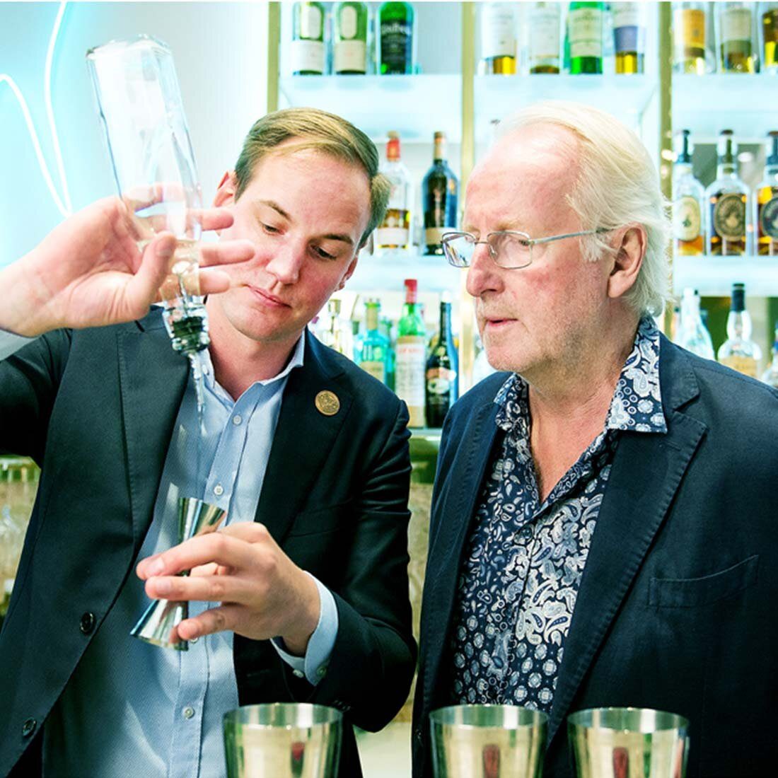 Bildet viser to menn, Eyvind Hellstrøm til høyre. Mannen til venstre heller klar væske fra en flaske og ned i et bartenders målebeger. I bakgrunnen skimtes hyller med ulike brennevin.