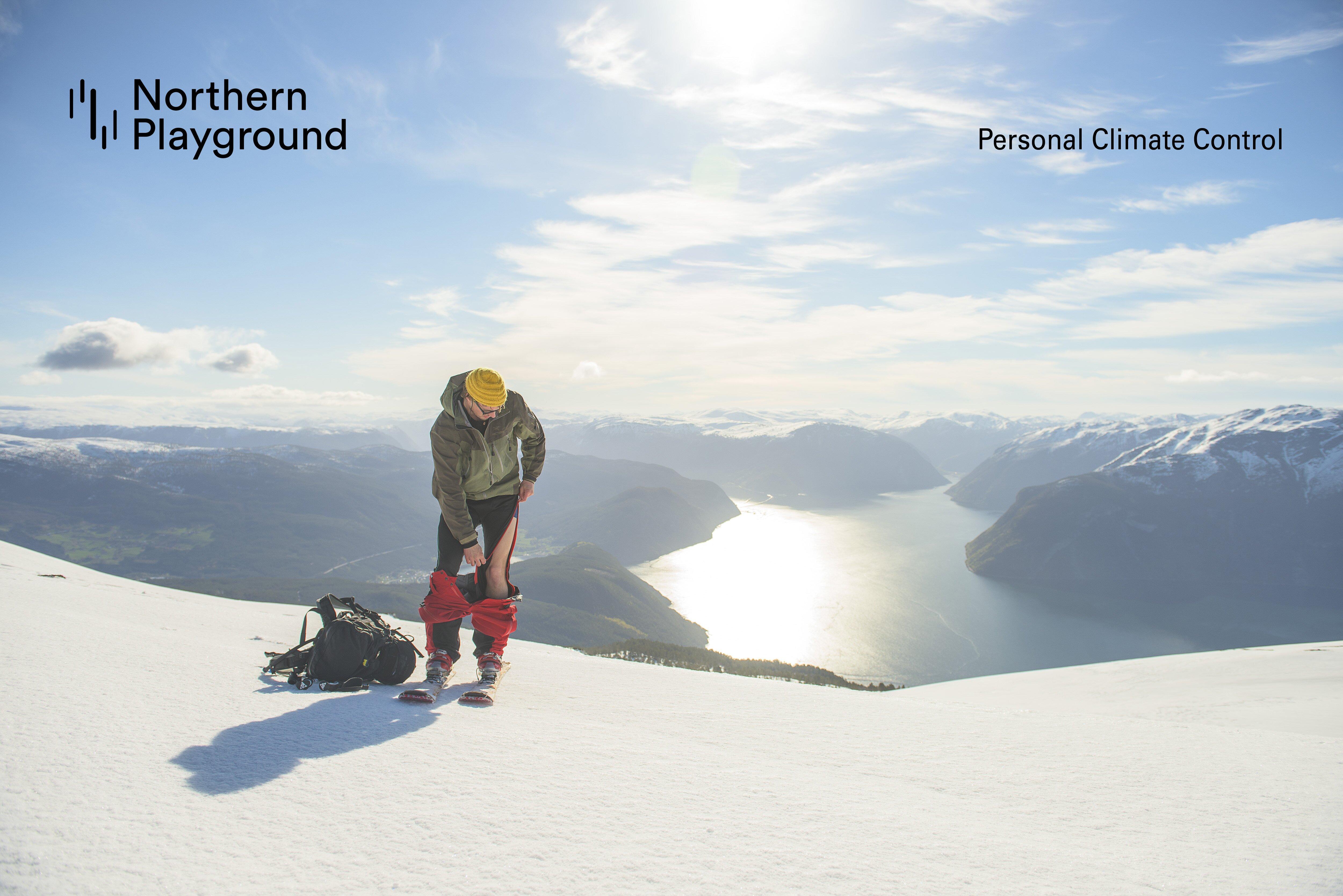Bildet viser en mann i turklær på en snødekt fjellrygg. Han har trukket ned skallbuksen og trekker opp en glidelås på ull-longsen under. Bak ham ses et vakkert fjordlandskap.