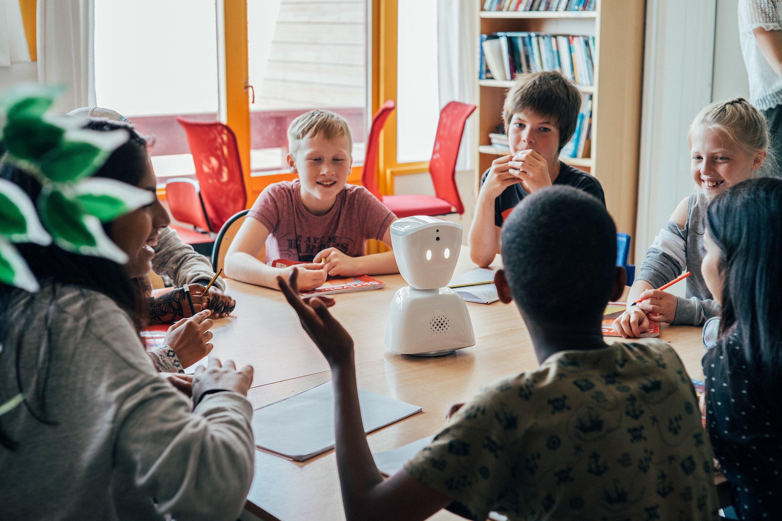 Foto av barn i skolemiljø. De sitter rundt et bord og har en samtale. I midten av bordet er AV1. En liten hvit robot – omkring 30 cm høy – med to lysende "øyne" og mikrofon/høytaler på "magen"