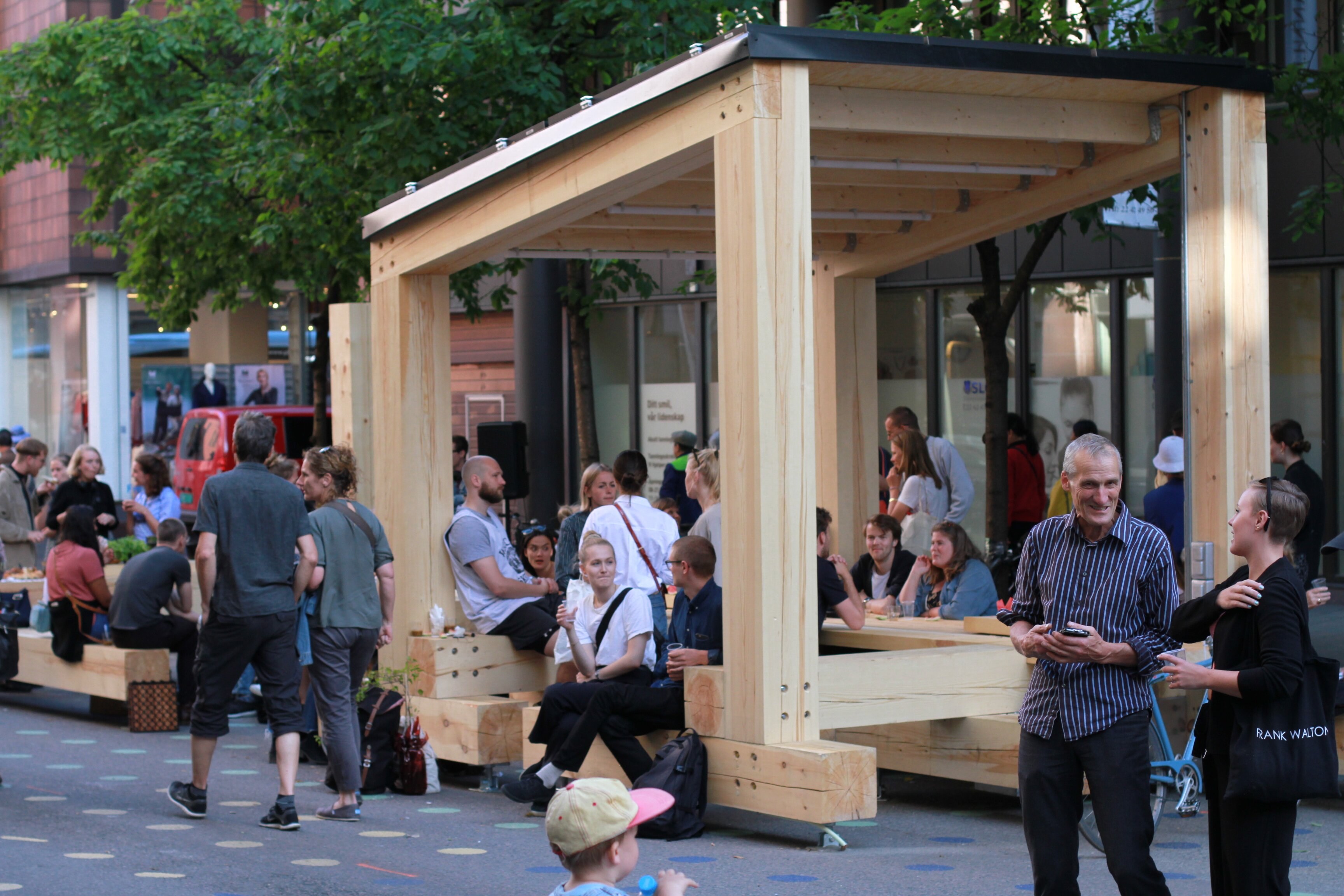 Et gatemiljø med yrende folkeliv. Sentralt i bildet står "Langbordet", et stort overbygget oppholdsmøbel.