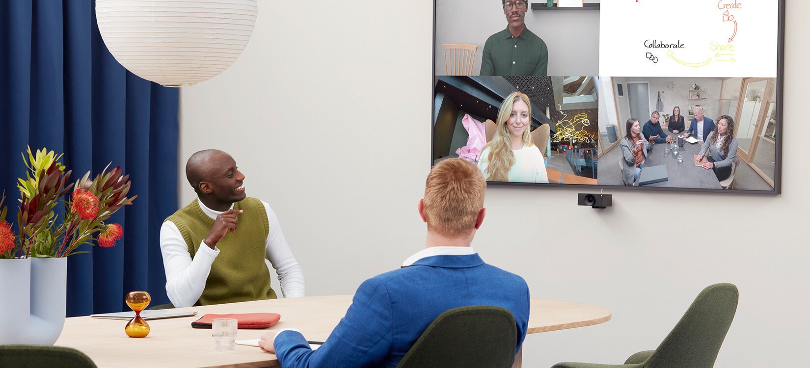 To personer i et møterom smiler mot en skjerm hvor vi ser andre møtedeltakere.