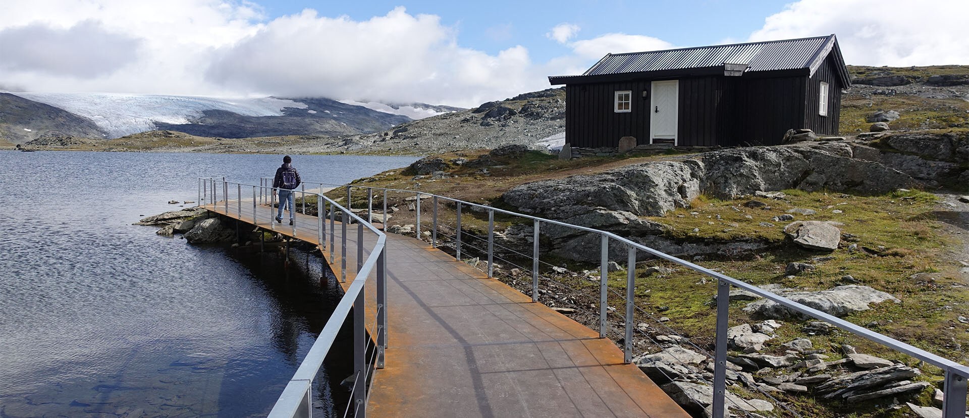 En person vandrer på en stålbro langs et vann. I bakgrunnen ses høyfjellslandskap og snødekte fjell.