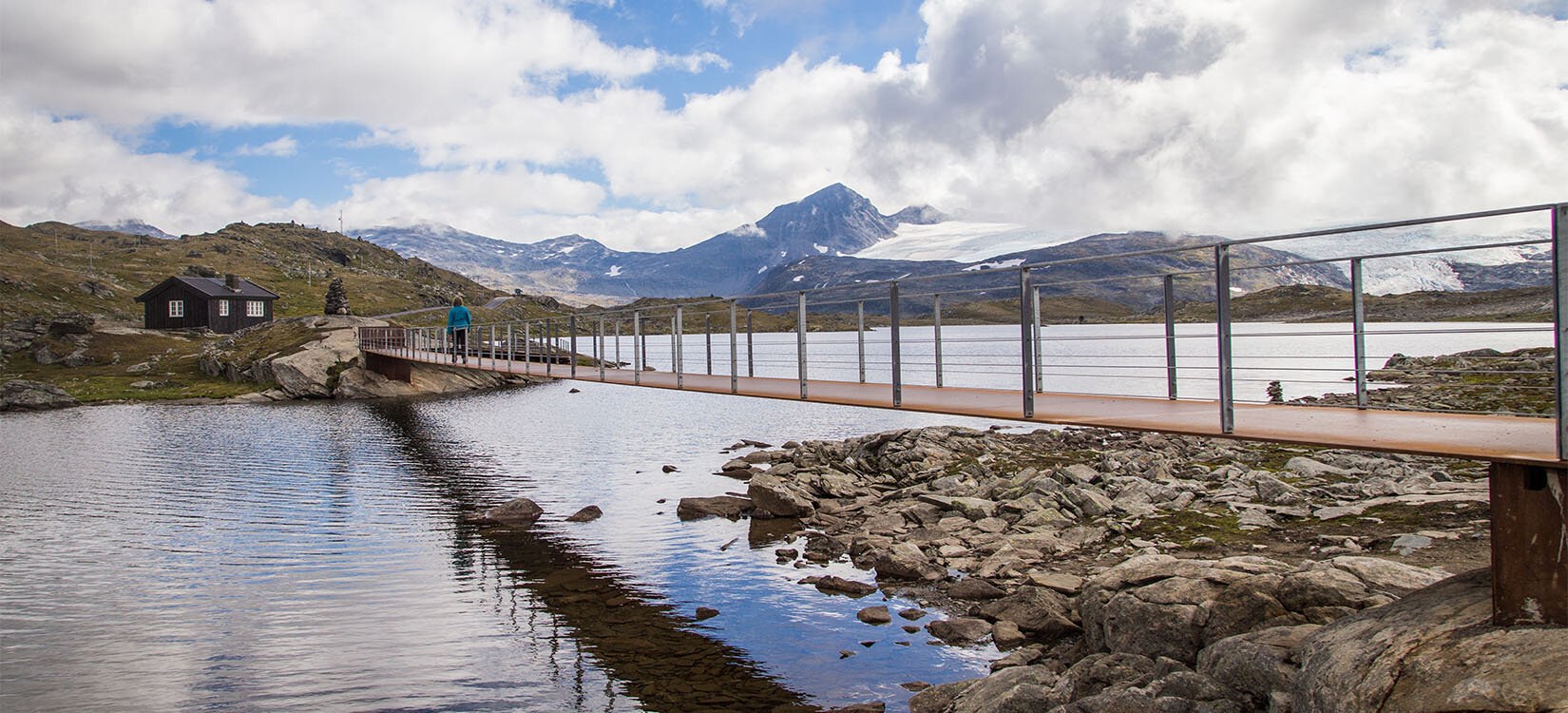 En bro strekker seg over et vann. I bakgrunnen ses høyfjellsnatur og fjelltopper.