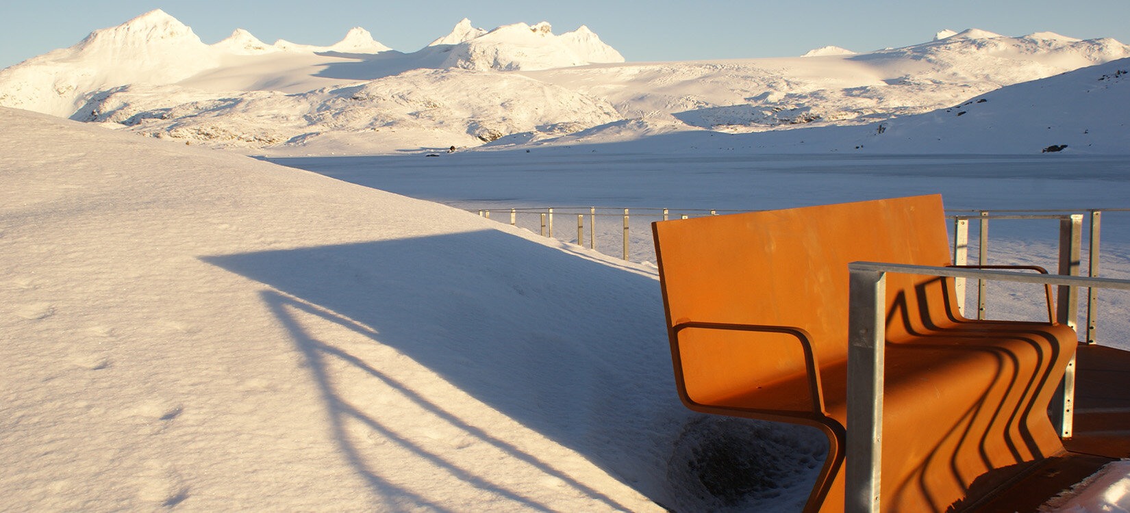 En benk i et snødekt landskap. I bakgrunnen ses en fjellkjede med karakteristiske spisse topper.