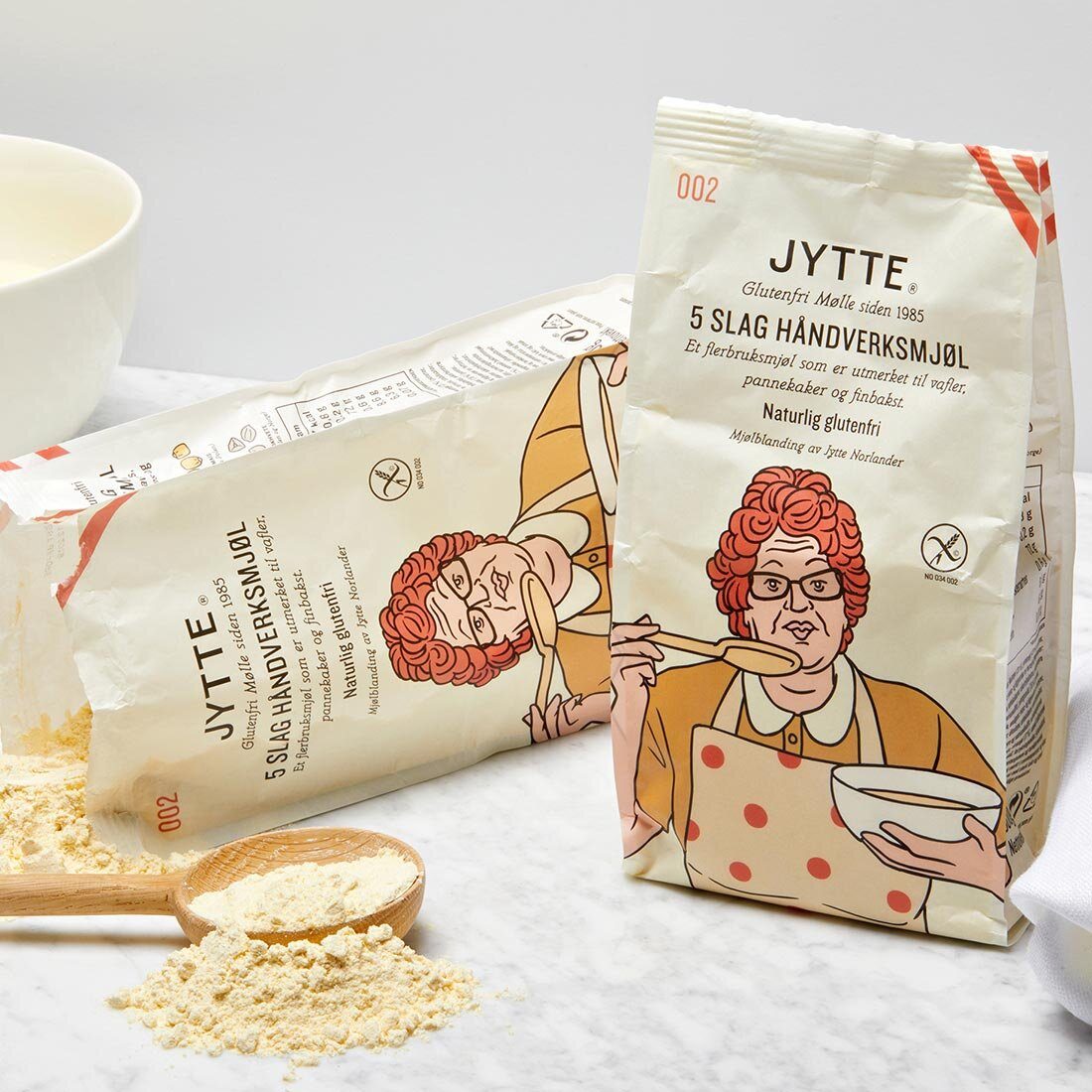 Bildet viser to pakker mel, den ene har veltet og lyst gult mel ligger utover en marmorplate. Emballasjen har en illustrasjon av en forklekledd eldre kvinne med krøllete oransje hår som holder en bakebolle i ene armen og i den andre: en skje mot munnen.