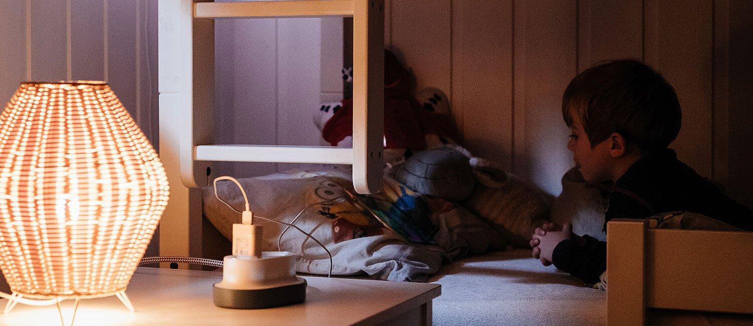 Bildet viser et barn som ligger i en dunkelt belyst seng. Fremst i bildet er en lader koblet i en hvit forgrener med svart base.