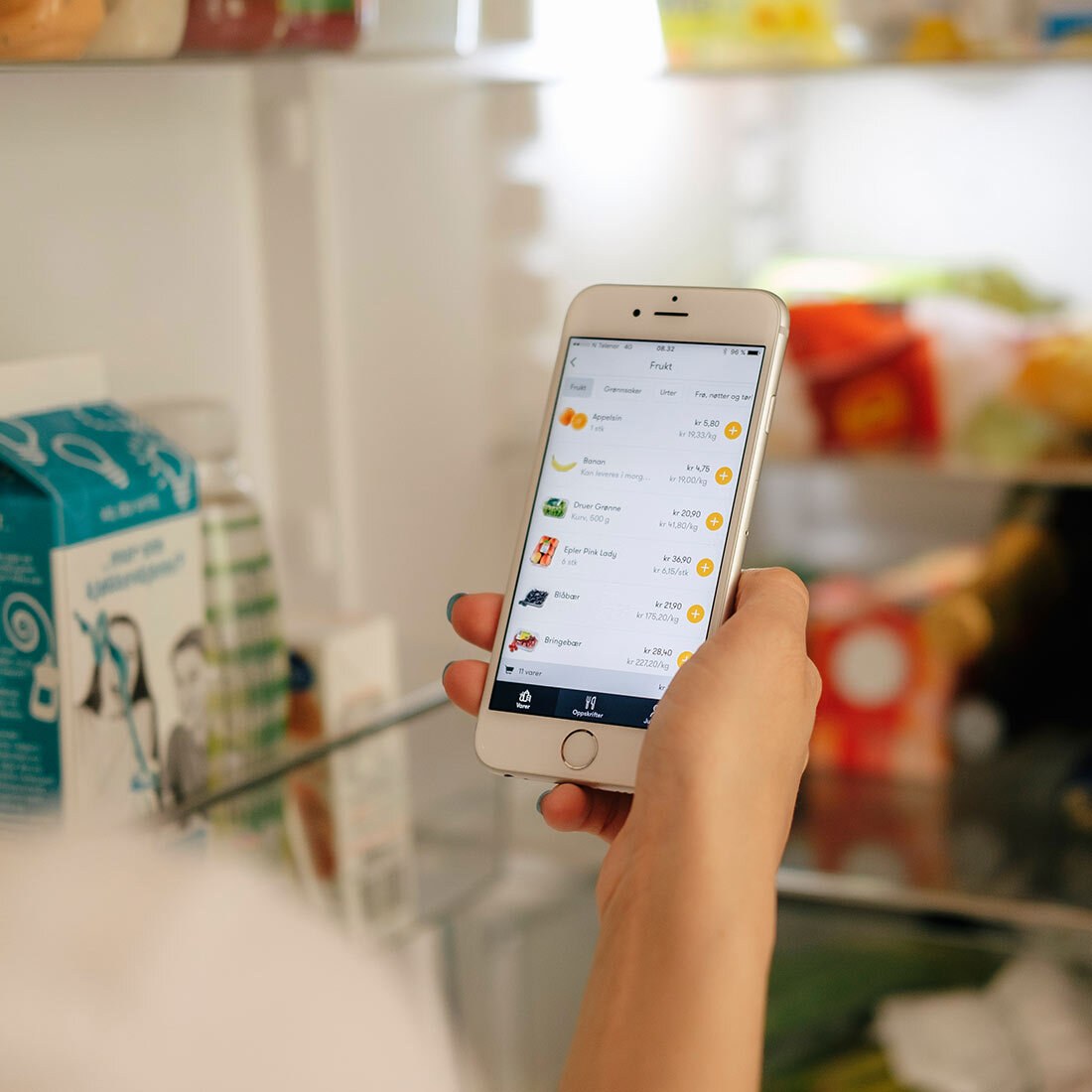 Vi ser en mobilskjerm med oversikt over ulike matvarer. Telefonen holdes av en kvinnehånd foran et åpent kjøleskap.