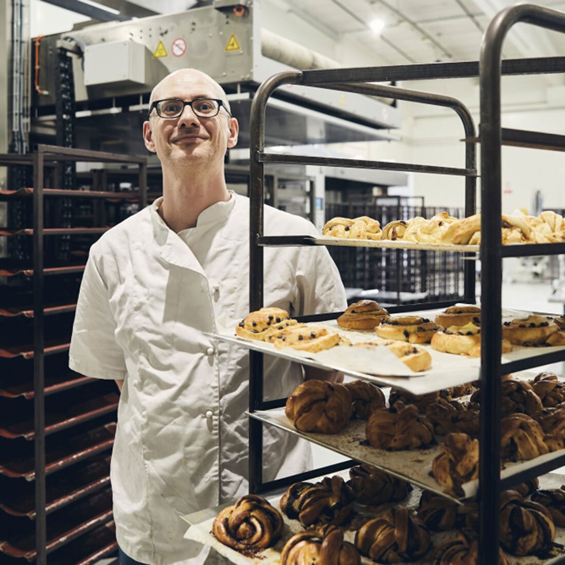 En mann kledd i hvit bakerskjorte står med rak rygg og smiler mot kamera, ved siden av et stativ med flere brett nybakte boller.