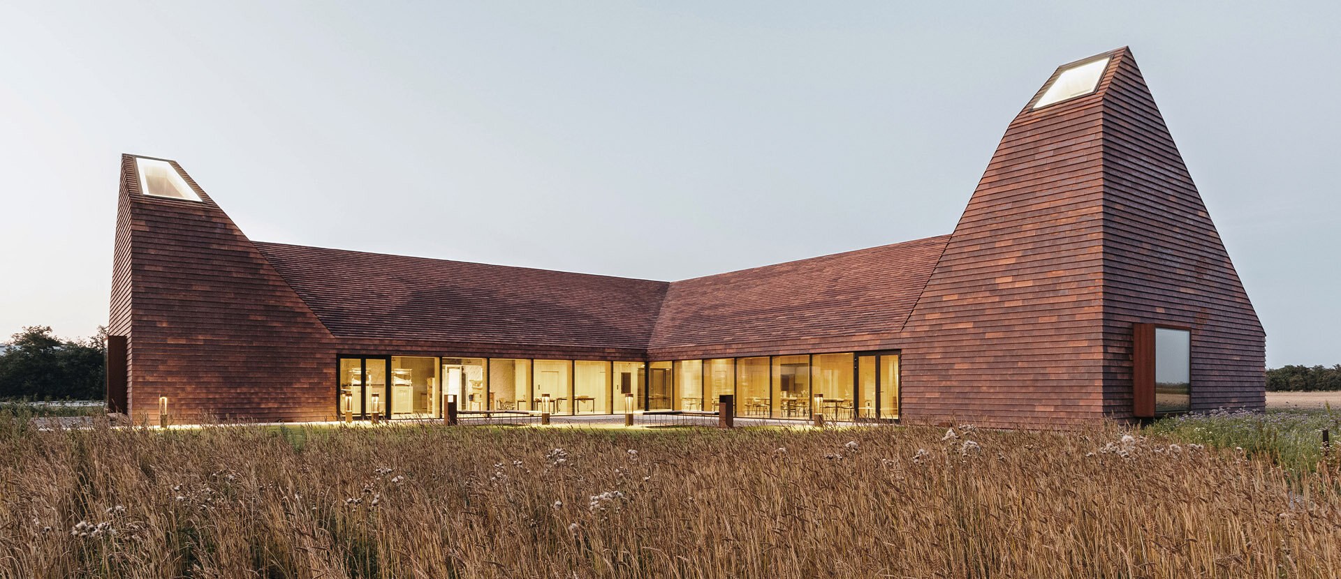 Foto av kornets hus: Et lavt bygg i en kornåker. Bygget er formet som en vinkel, med to pyramide-formede tårn i enden av hver fløy. Fasade og tak er kledt i liggende panel i rødbrune nyanser.