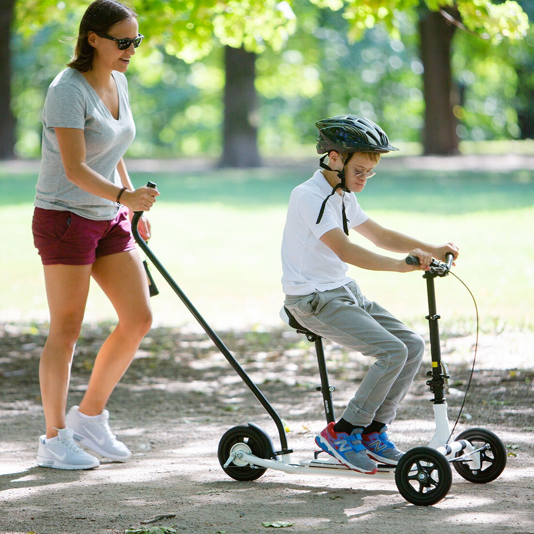 Bildet viser en ung gutt på sparkesykkel. Han sitter på et sete og holder i styret, mens en kvinne går bak og holder i et håndtak som er festet til sykkelens bakdel.
