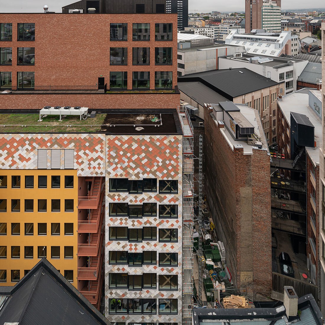 Bygningsfasader i et urbant bymiljø. Ett av byggene utmerker seg med fargbruk i fasaden: gult, flerfargede plater og terracotta.