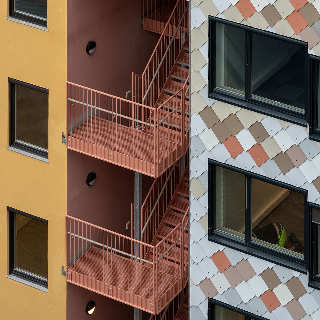 Et halvnært bilde som viser farger og detaljering i en fasade. Gulmalt bygg, mot terracottafarget trappegang, mot en platekledt fasade i dempede nyanser av blå, brunt og rødt.