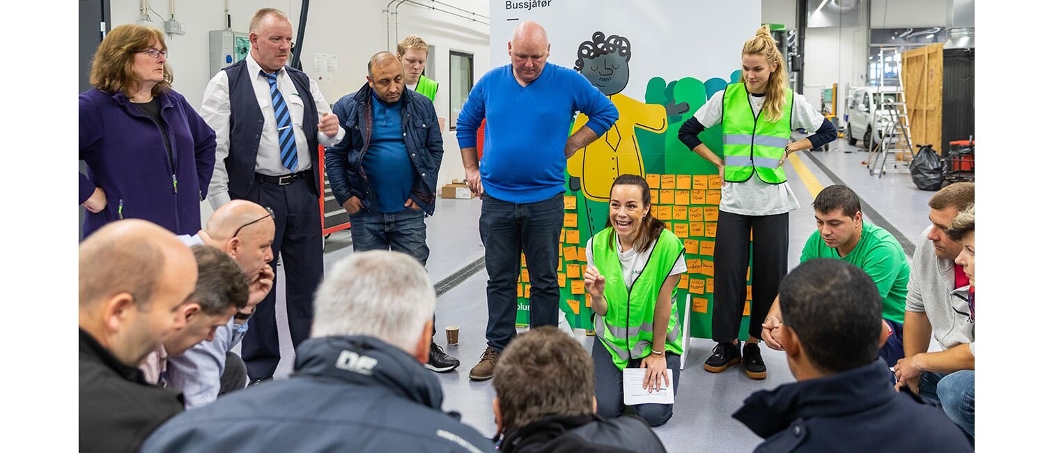 Bildet viser en gruppe mennesker som står rundt en kvinne i grønn refleksvest som sitter på knærne på gulvet og prater til forsamlingen.