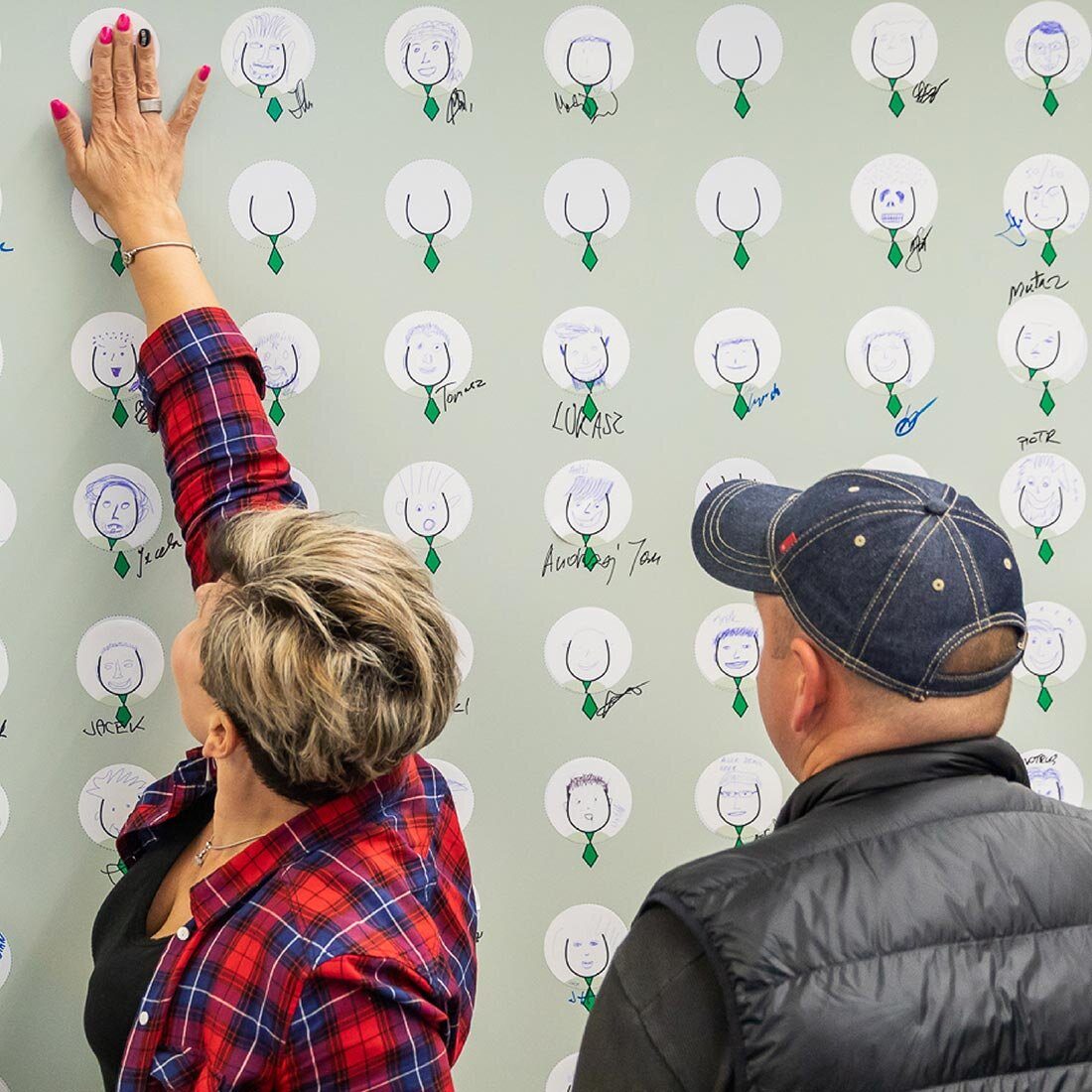 En kvinne strekker seg for å ta på en tegning på veggen. Tegningen er av det sammen hodet med et grønt slips. Noen av hodene har fått håndtegnede ansikter og navn.