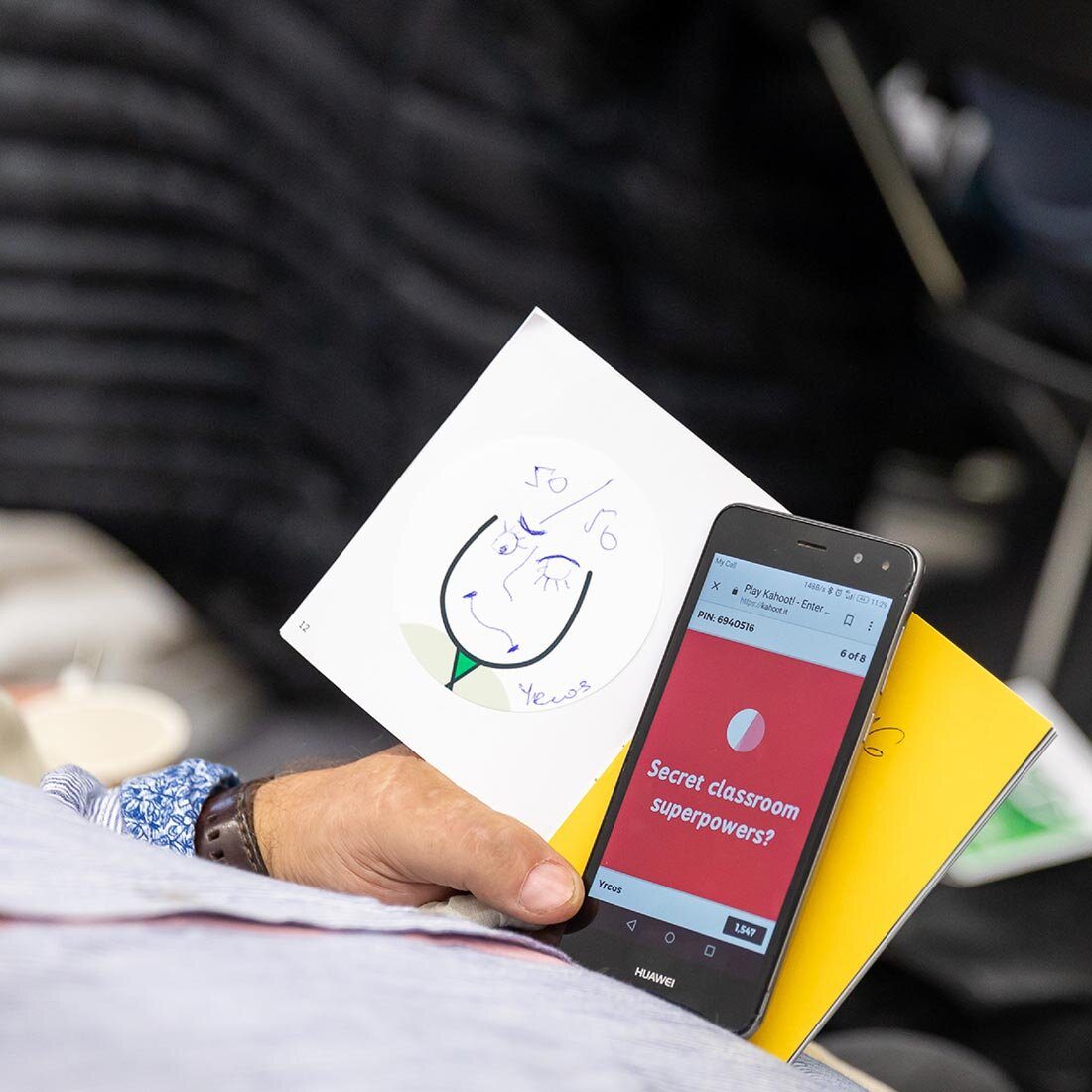 Et nærbilde av en manns hånd som holder en brosjyre og en mobiltelefon. Brosjyren har en illustrasjon av et hode med grønt slips og håndtegnet ansikt. På telefonen har han kahoot.