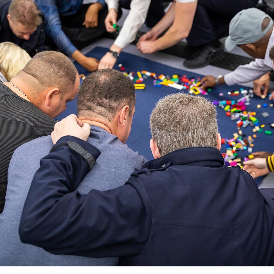 Bildet viser ryggene på tre menn som sammen med flere sitter rundt det som ser ut som legeklosser på gulvet. Flere strekker seg etter klosser. En av mennene holder vennskapelig på ryggen til sidemannen.