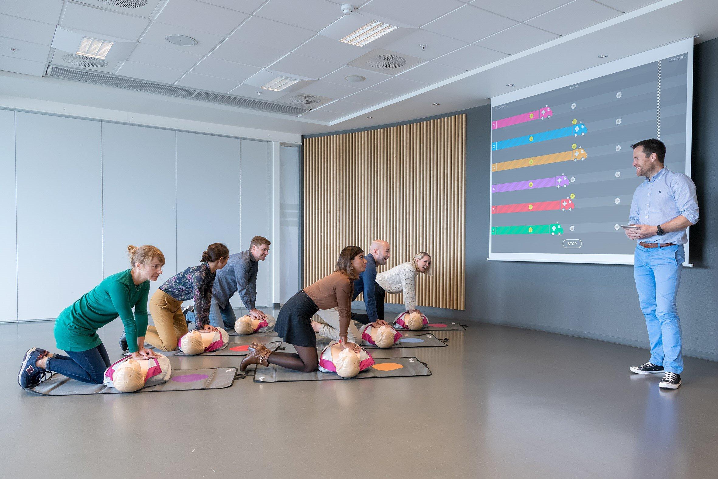 Bildet viser et klasserom med en instruktør som står, mens seks voksne kneler på gulvet og gir kompresjoner til hver sin Anne-dukke. På et lerret vises de ulike deltakernes progresjon i typisk spill-uttrykk med sterke farger og et barnlig uttrykk.