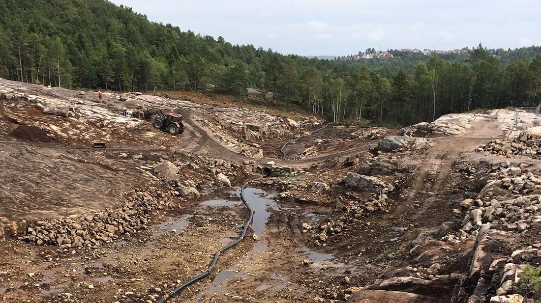 Foto av område, omkranset av en skog, som er preget av aktivitet: ingen vegetasjon, kun jord, stein, hjulspor, jordveier og dype grøfter og rør.